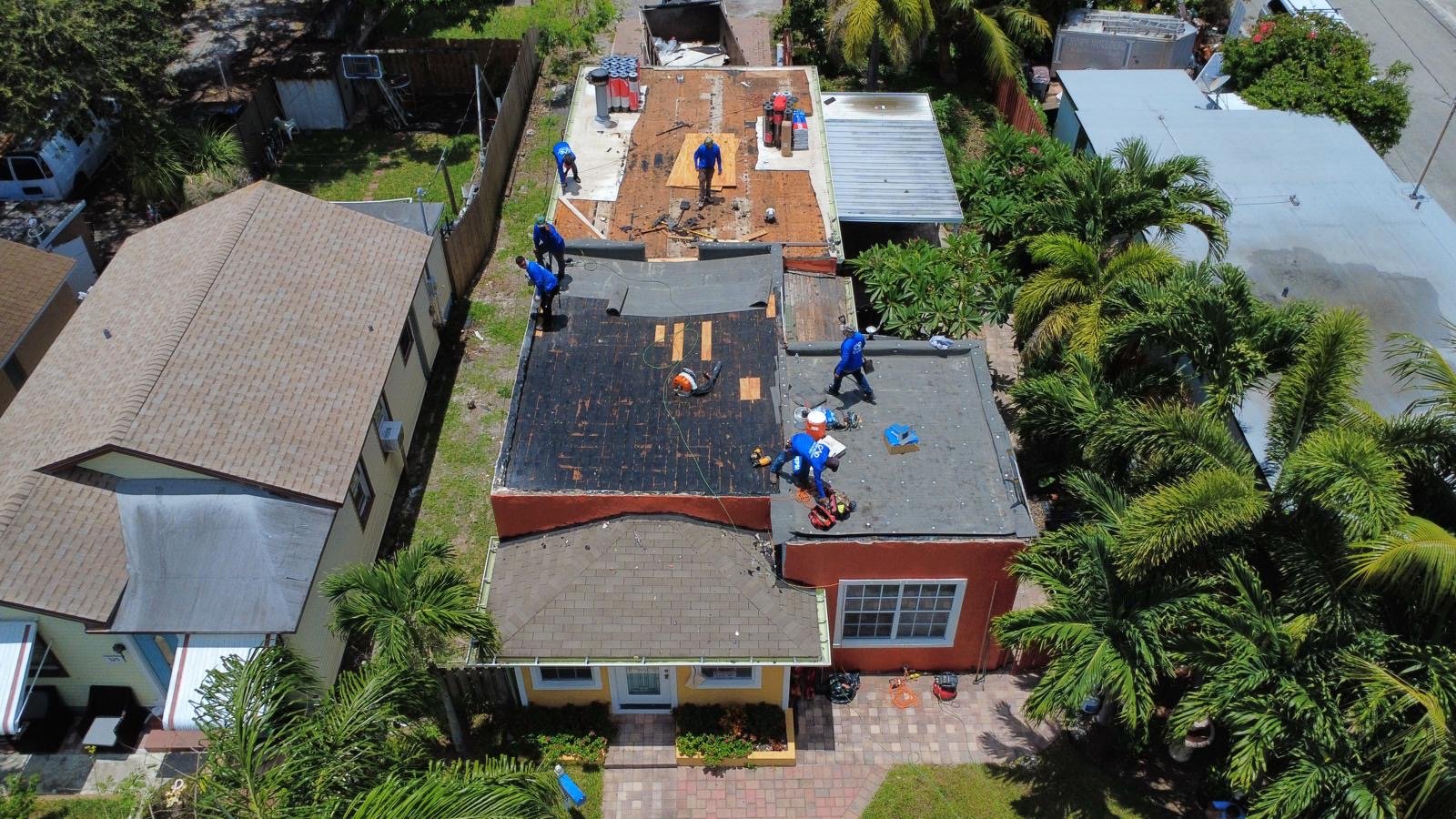 Aerial view of workers repairing a flat roof on a residential building, with some sections of the roof already stripped and others being worked on, surrounded by neighboring houses and lush green trees.