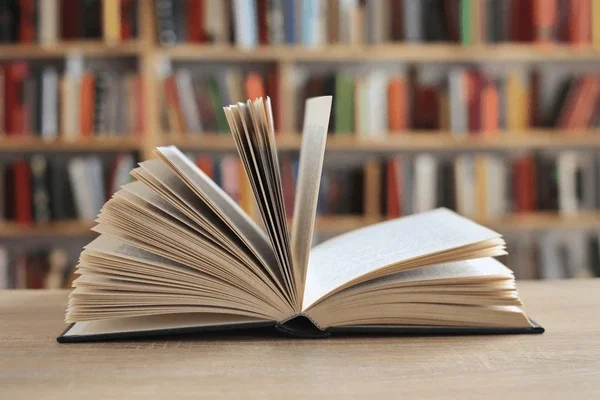 Open book on a table in front of a bookshelf with many books in the background.