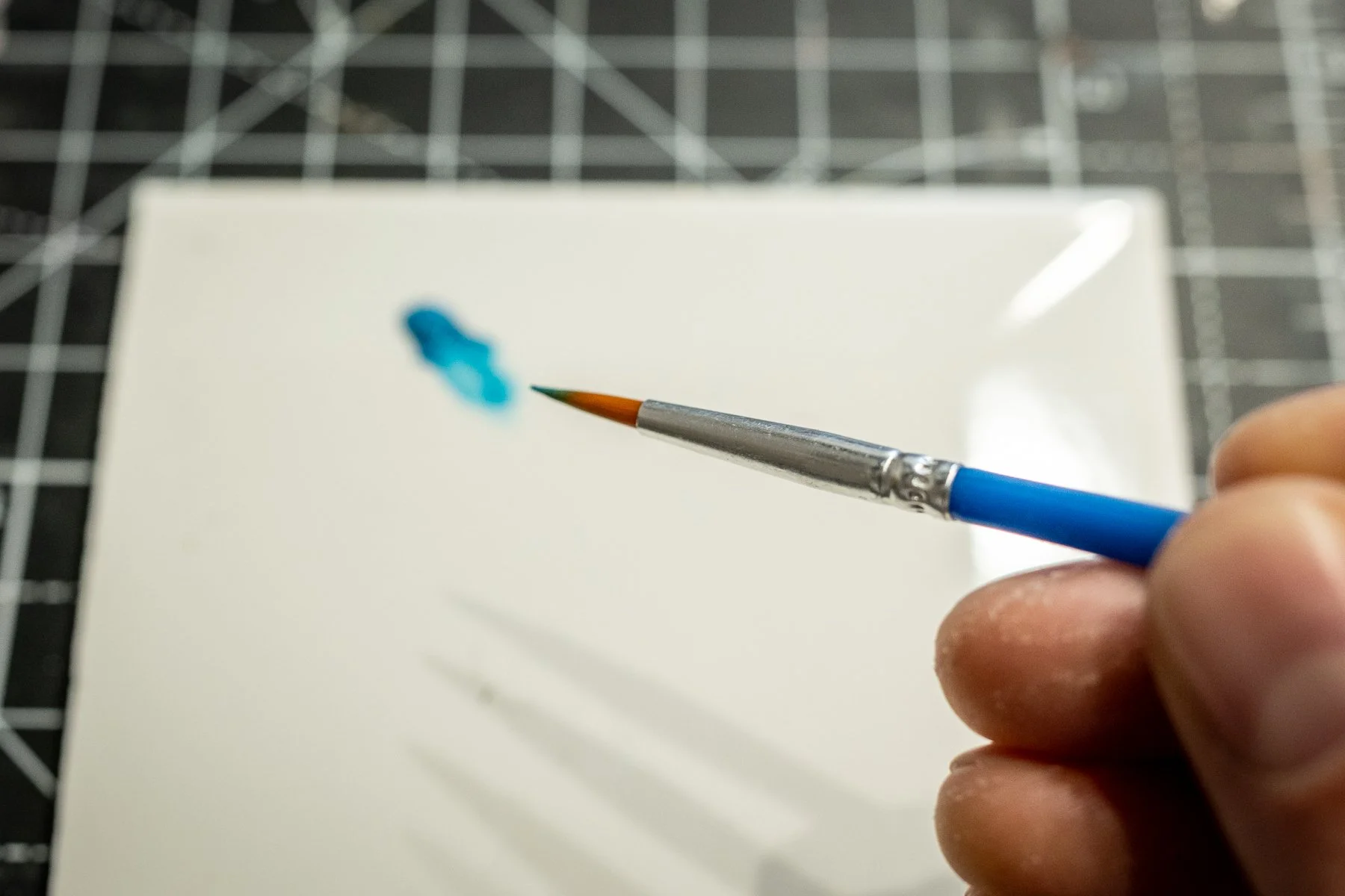 A hand holding a paintbrush with blue and green paint, painting on a blank white canvas.