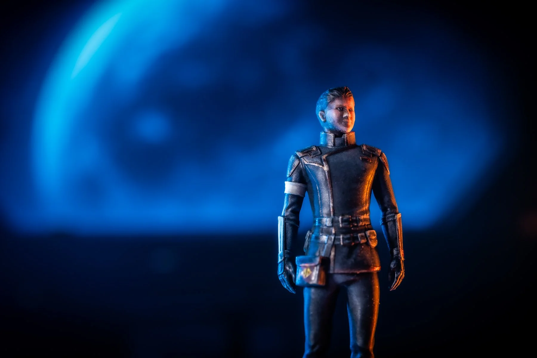 A female action figure standing on a dark surface, lit by blue and orange lighting, with a blurred blue background.