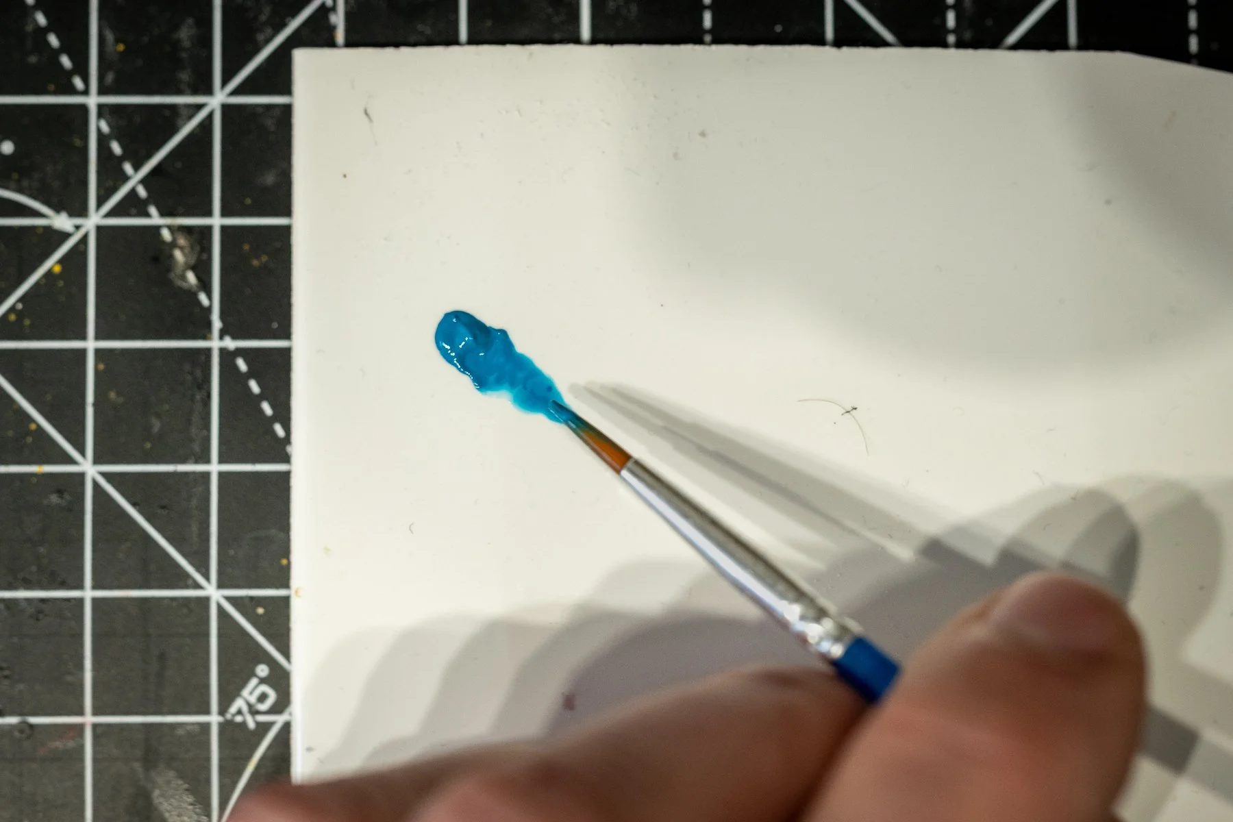 Close-up of a paintbrush with blue paint on the tip, held over a white surface, with a black cutting mat marked with grid lines in the background.