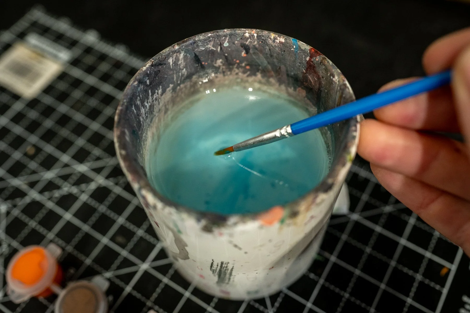 Hand stirring blue liquid paint in a metal container with a fine-tipped brush, on a grid-patterned black work surface.