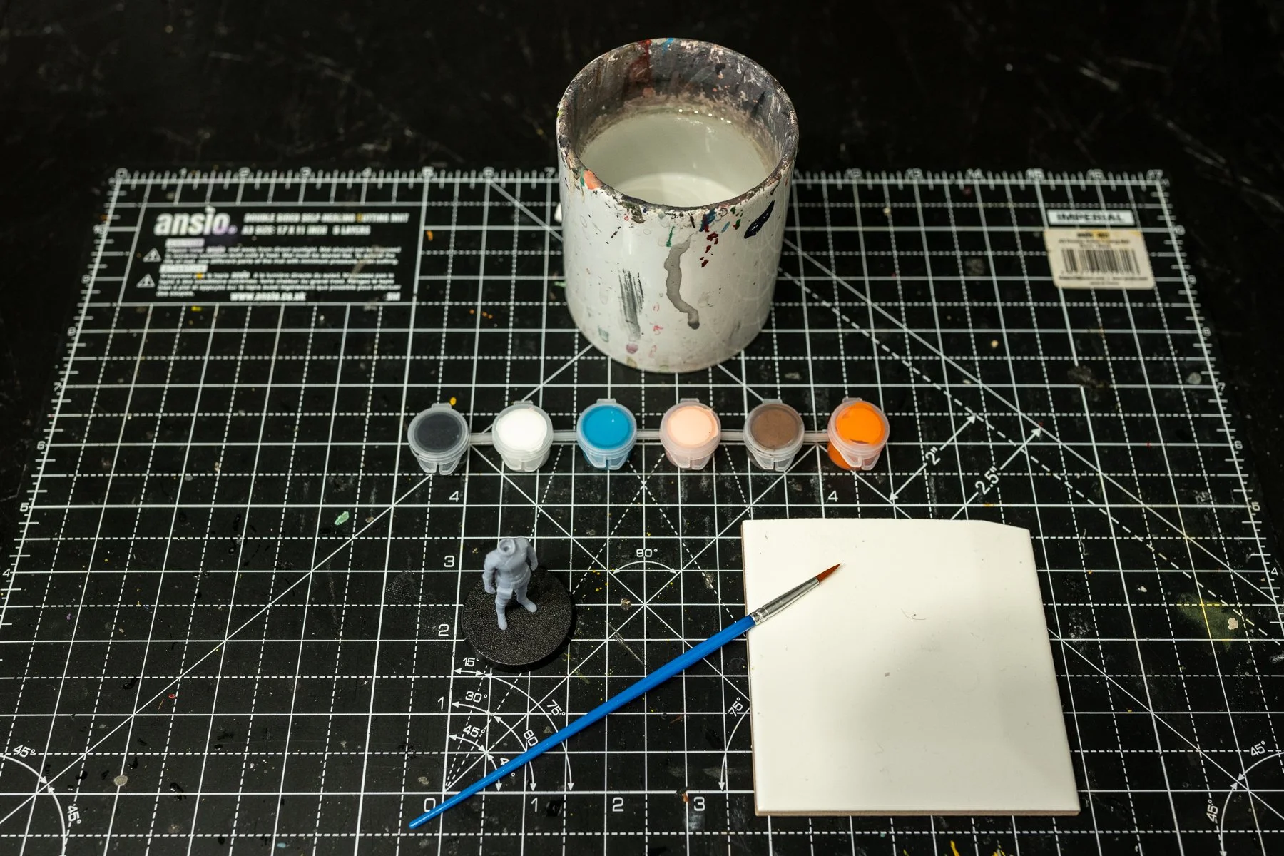 Art supplies and miniatures on a black cutting mat. Contains a paint cup, six paint pots in gray, white, blue, peach, brown, and orange, a small astronaut figurine on a black base, a paintbrush, and a white notepad.
