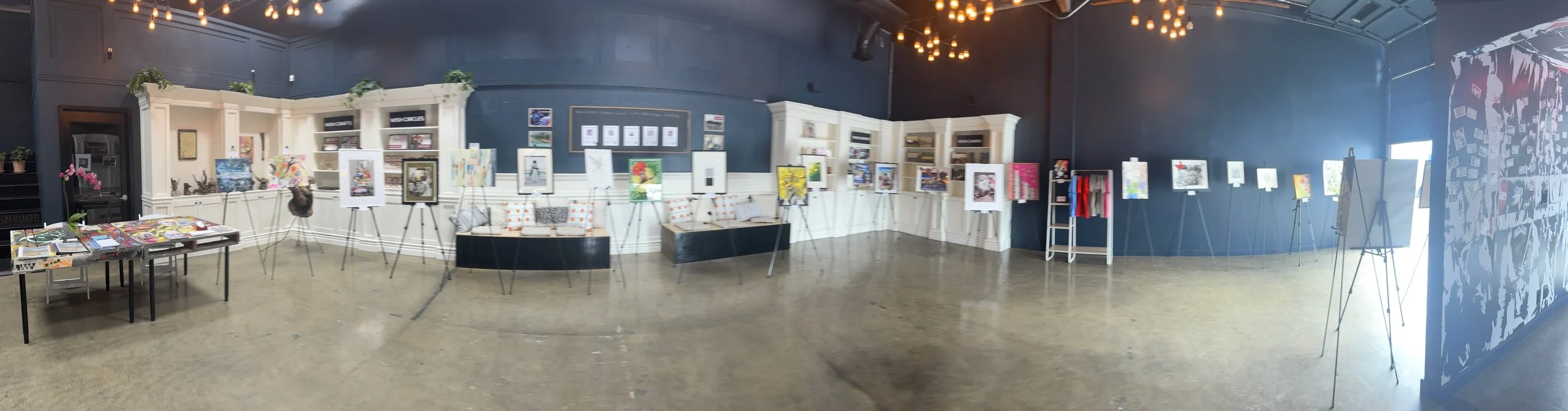 An art gallery with paintings on display on easels and shelves, and tables with art books, in a room with dark blue walls and warm lighting.
