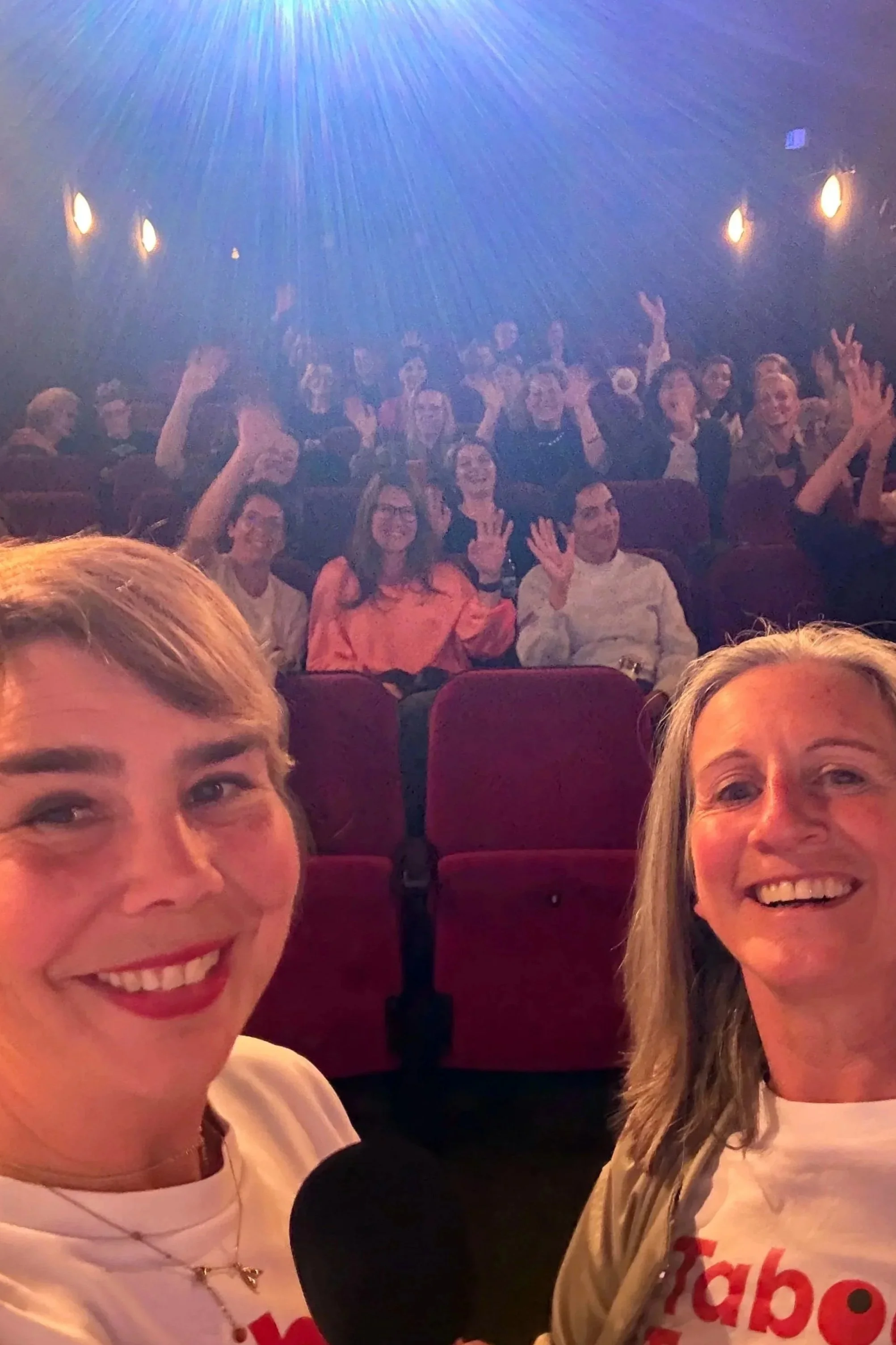 Zwei lachende Frauen in einem dunklen Theater, die für ein Foto posieren, während im Hintergrund eine Gruppe Menschen fröhlich winkt.