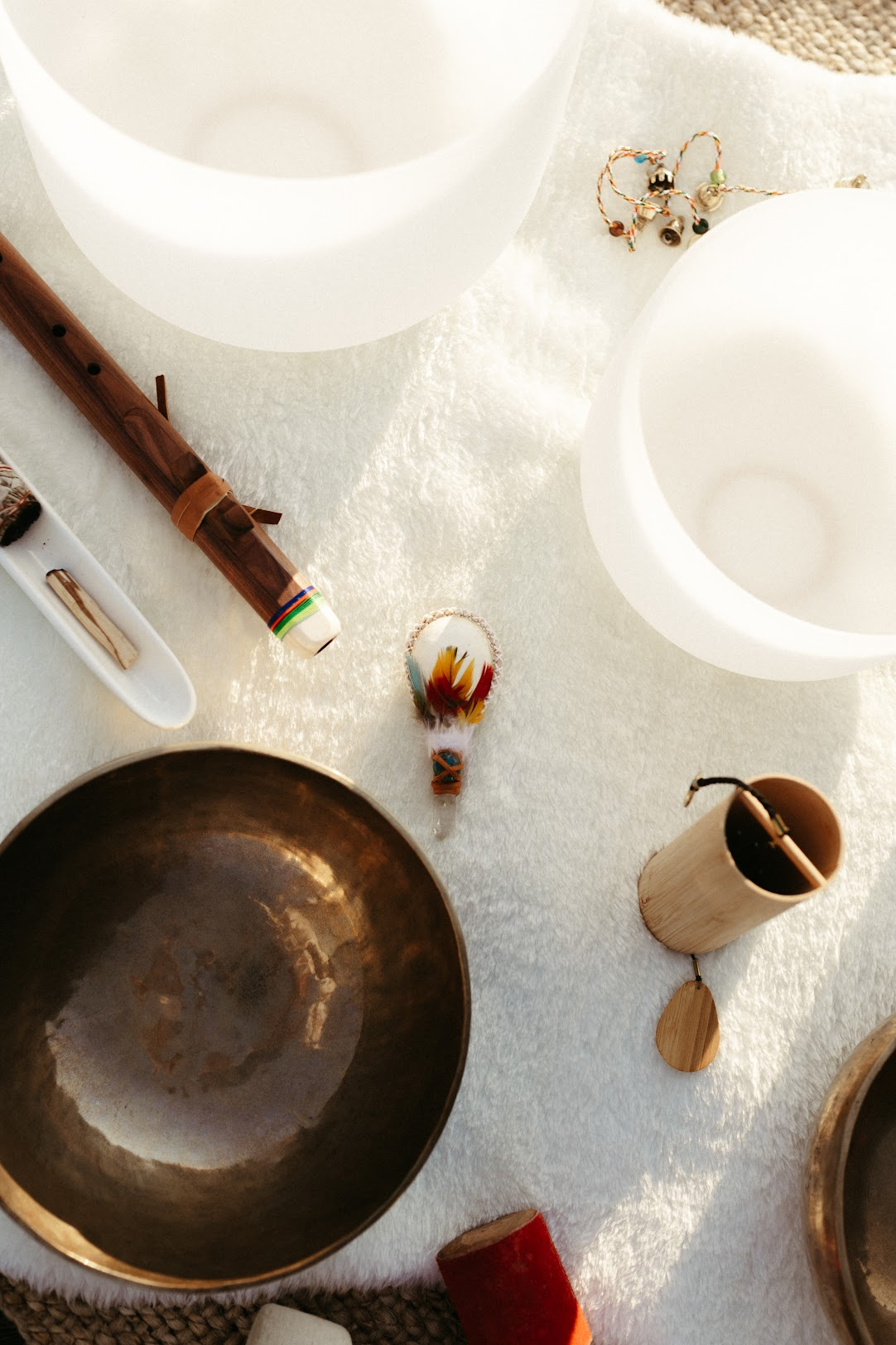 A collection of spiritual and musical objects on a white surface, including Tibetan singing bowls, a flute, a feathered drum, incense holder, and jewelry, with two large white candles in the background.
