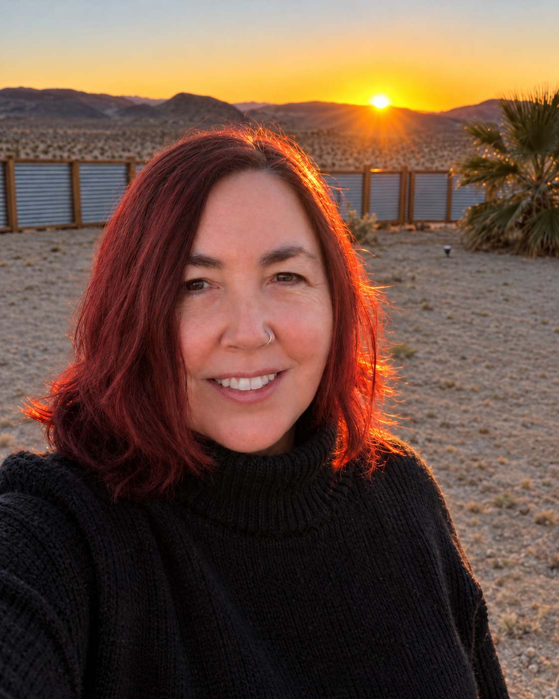 A woman with red hair and a nose ring taking a selfie in a desert landscape at sunset, with a fence and mountains in the background.
