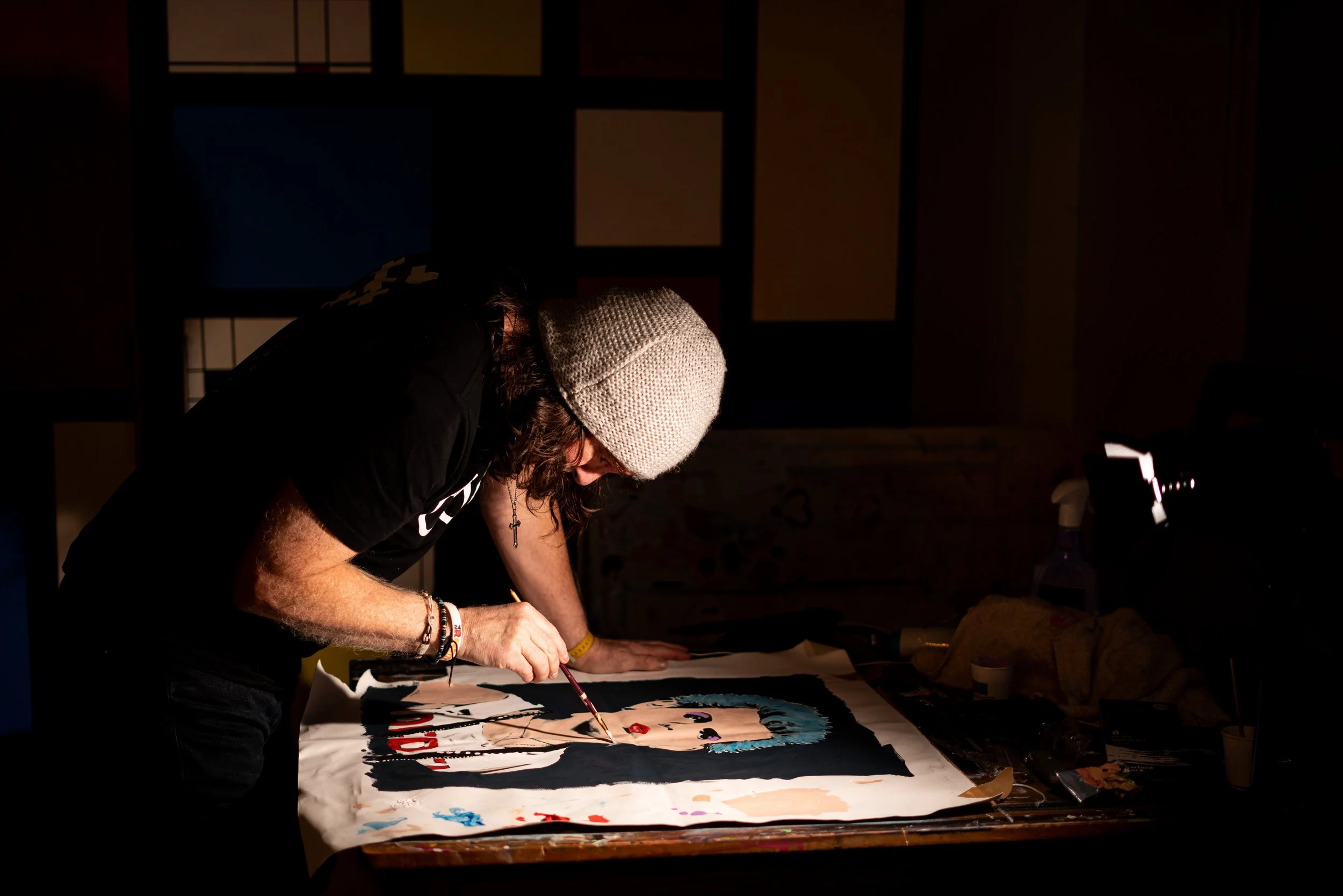 An artist wearing a gray knit beanie and black t-shirt working on a colorful portrait painting on a large sheet of paper, in a dimly lit room.