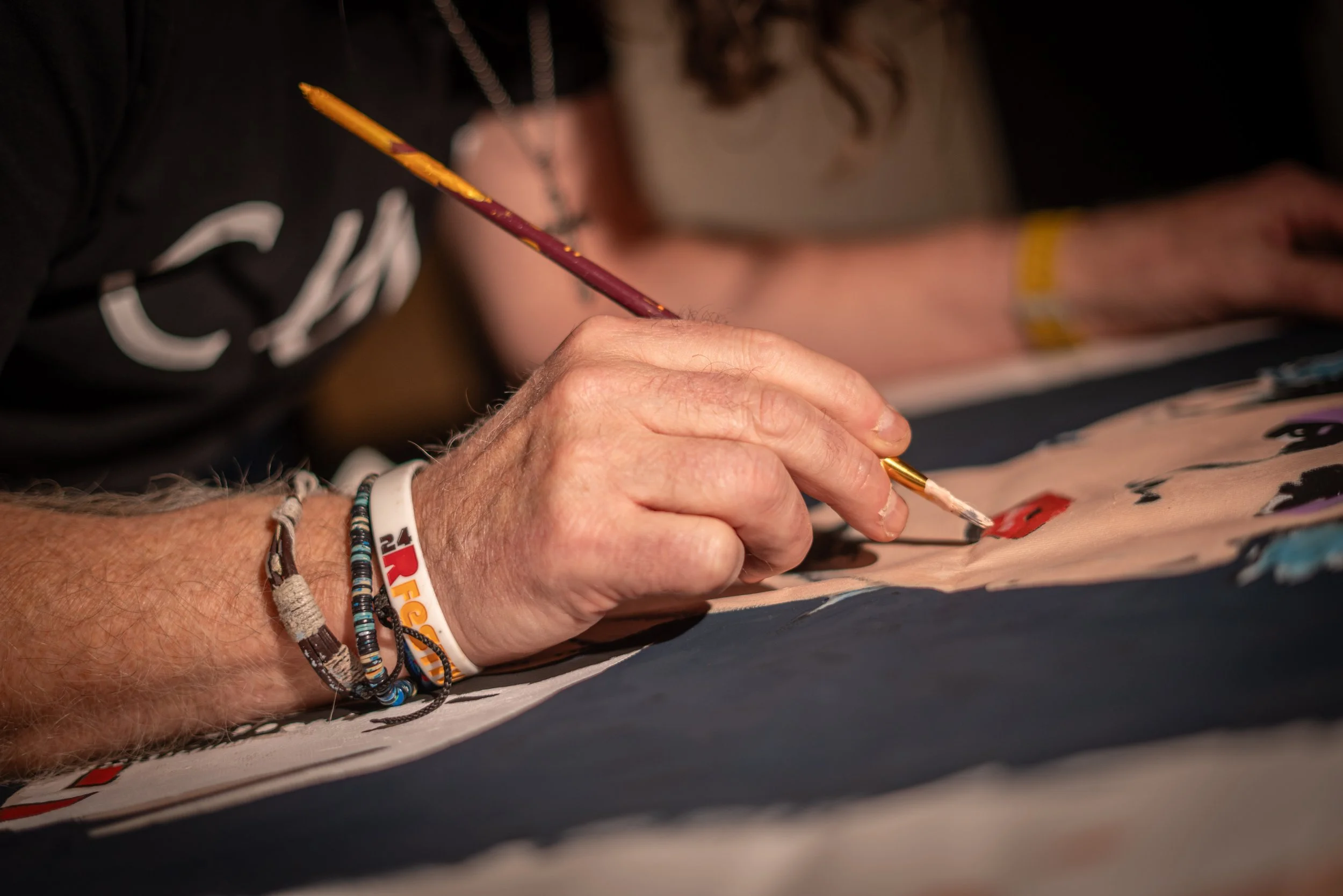Close-up of a person painting on a canvas with a fine brush. The individual has a wristband and bracelets, and there are other people in the background.