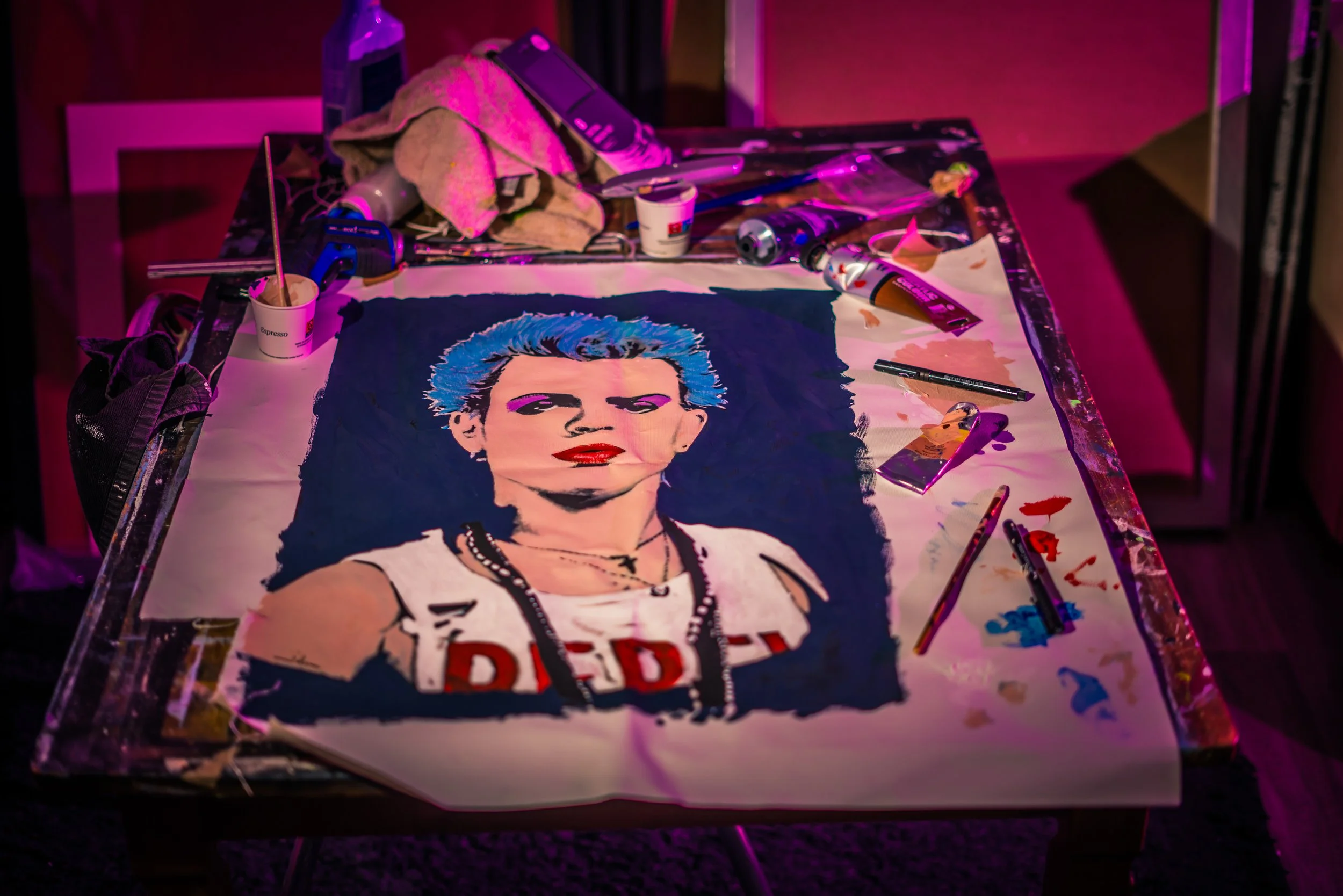 A portrait of a person with blue hair, red lips, and a septum piercing, painted on canvas supplies scattered around, on a table with pink lighting.