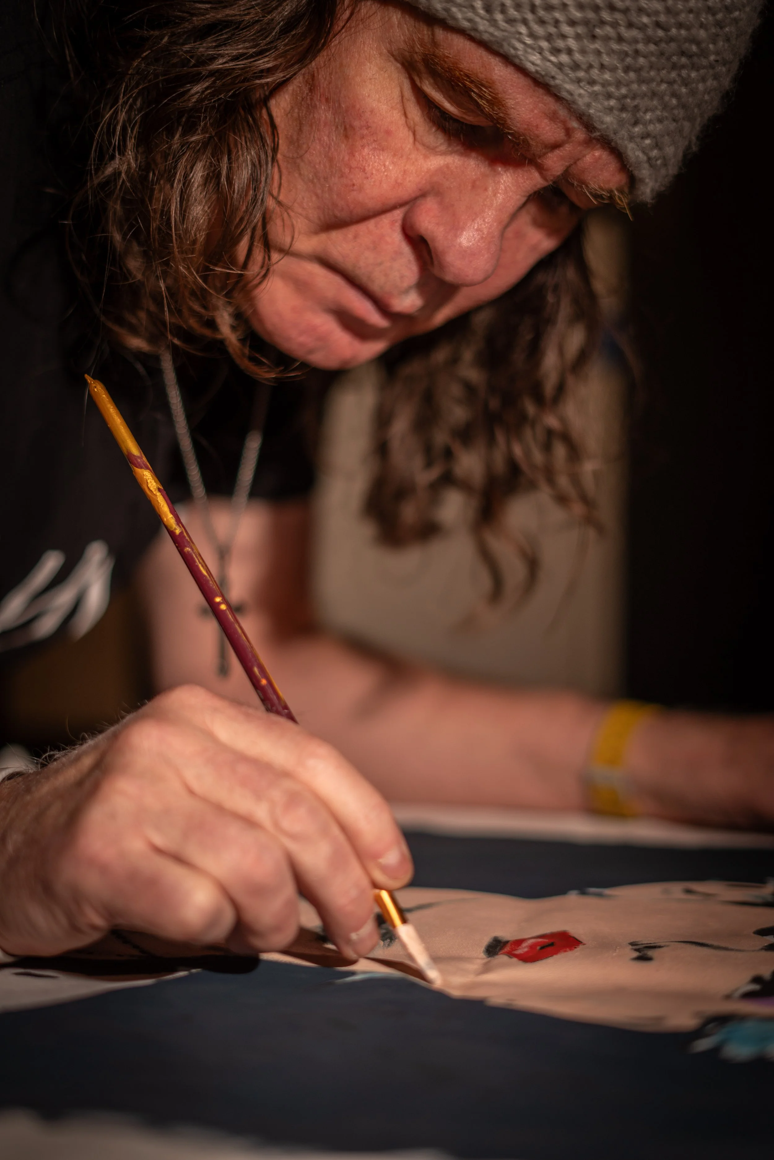 A person with long hair wearing a gray beanie and black shirt painting on a canvas with a brush.