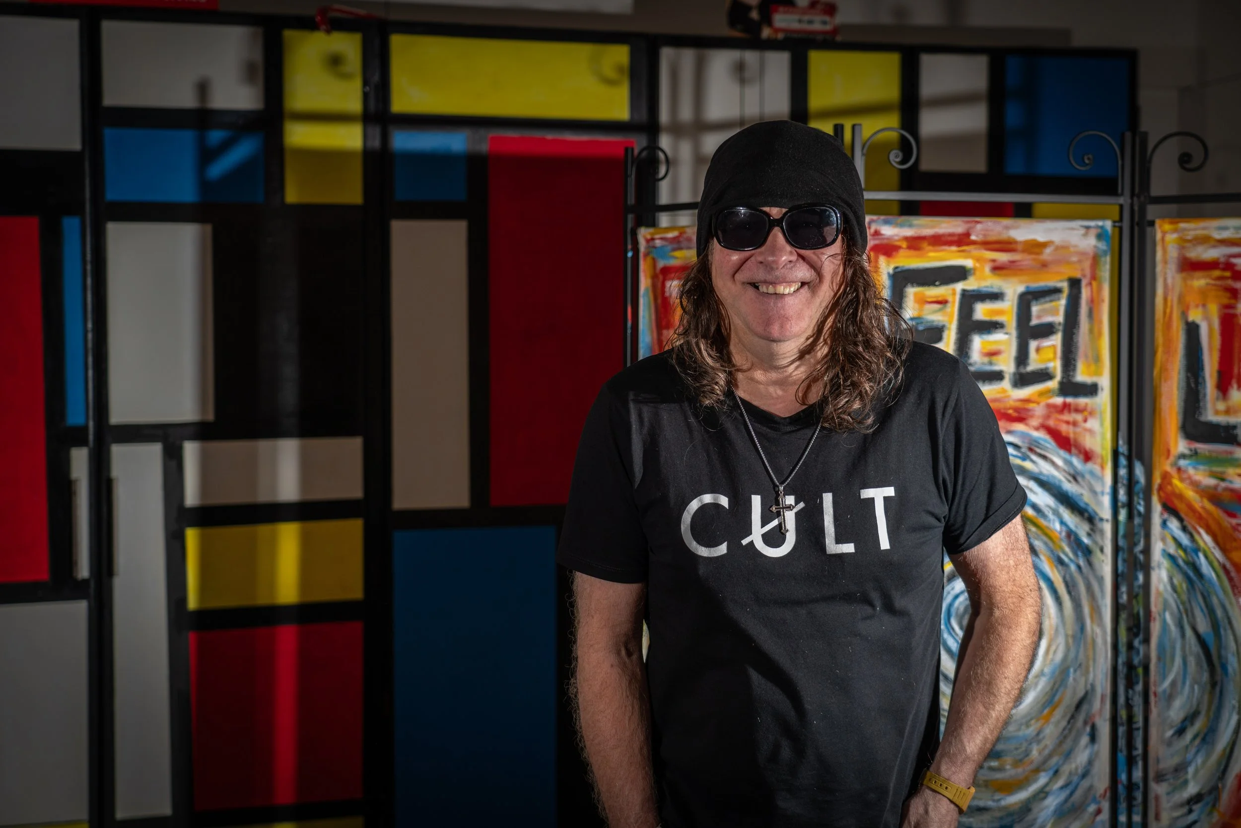 Smiling man with long hair, wearing sunglasses, a black beanie, a black T-shirt with "CULT" written on it, and a cross necklace, standing in front of colorful abstract art panels.