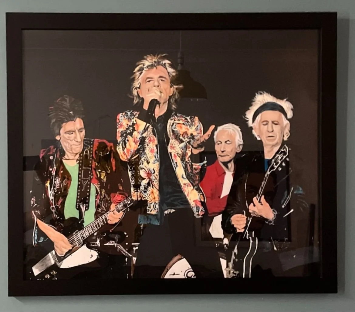 A framed photo of a musical band performing on stage, with four members, three playing guitars and one singing into a microphone, dressed in colorful and eclectic clothing.