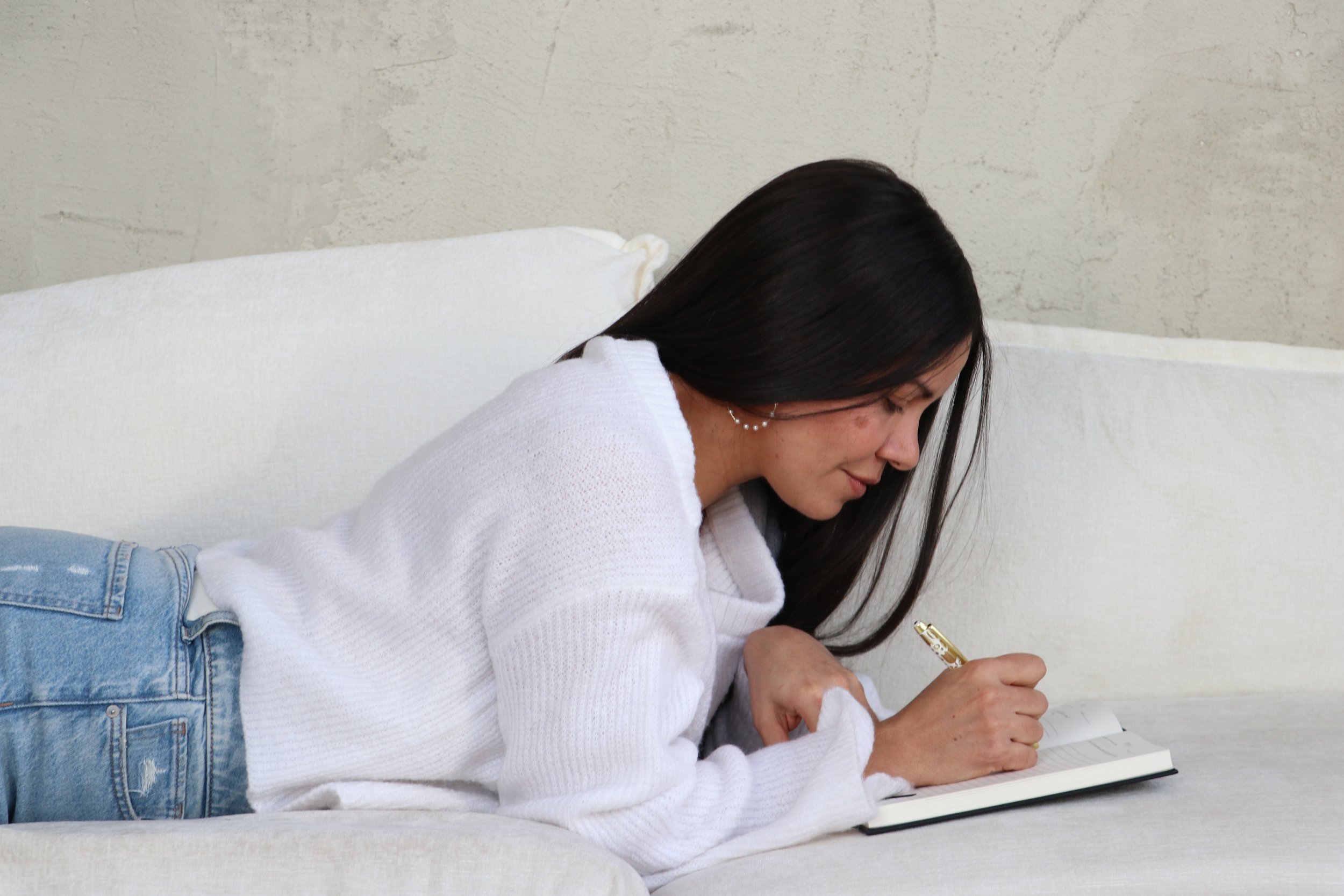 Mujer joven acostada en un sofá, escribiendo en un cuaderno con bolígrafo dorado.