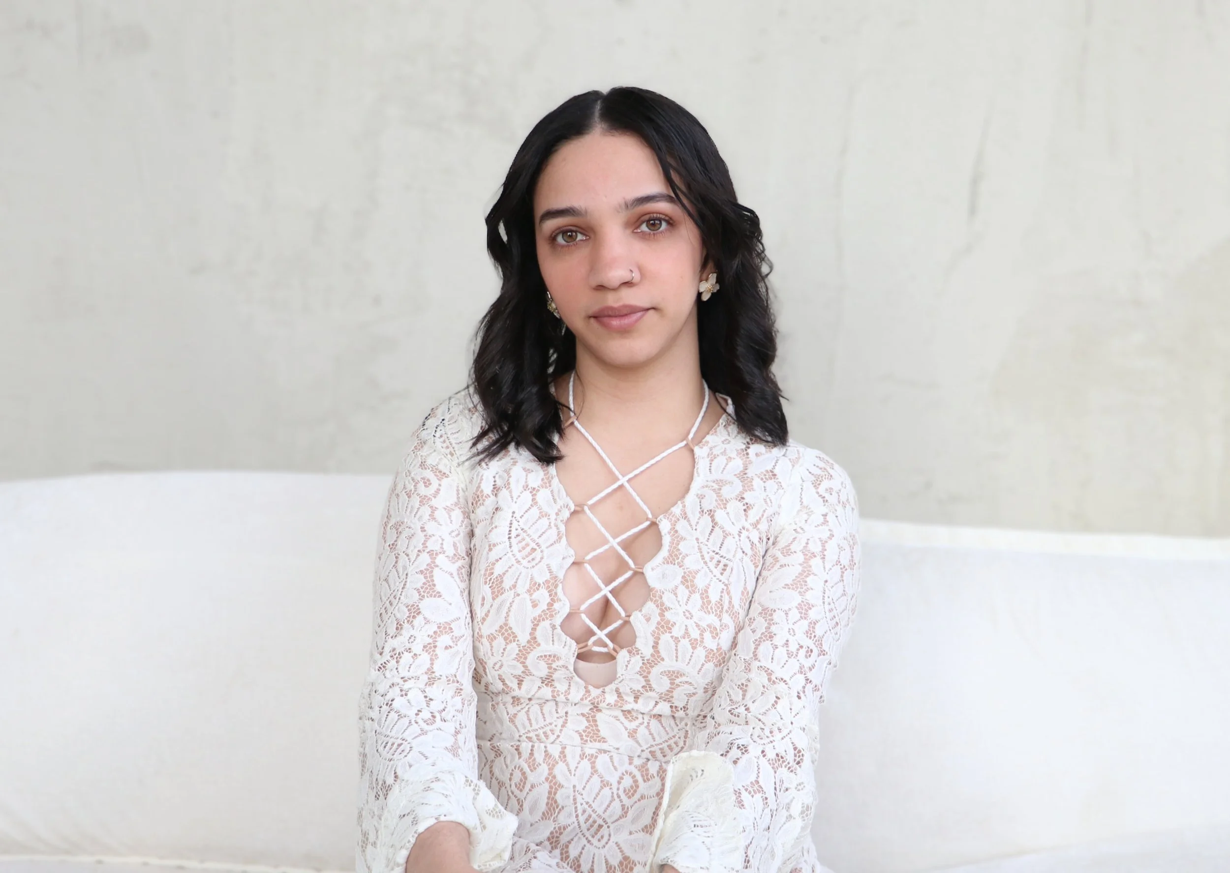 Mujer joven con cabello rizado negro, vestida con blusa de encaje blanca con diseño de cruces en el pecho, frente a fondo de pared clara.