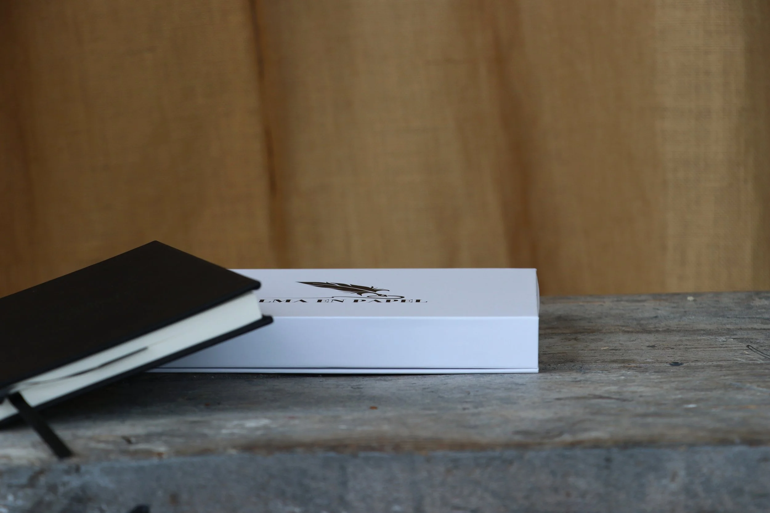 Un cuaderno negro con banda elástica y un libro blanco con logo de una pluma negra en la portada, ambos sobre una mesa de madera envejecida.
