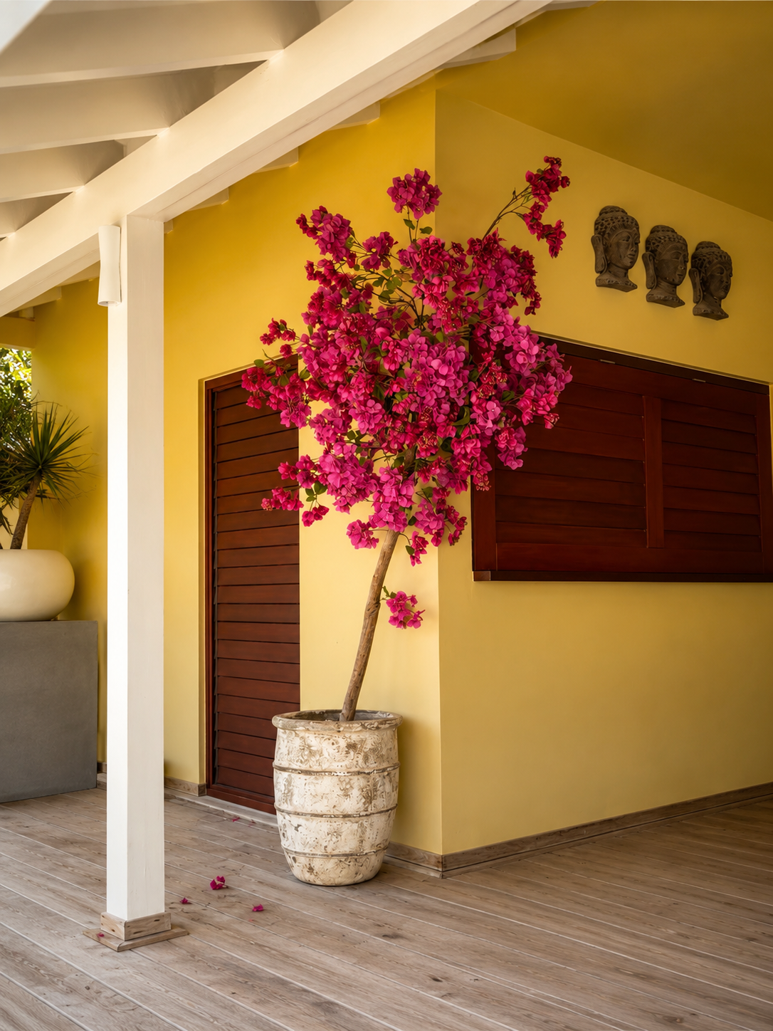 Indoor scene with a potted pink flowering plant, yellow wall, wooden shutter, decorative masks, and wooden flooring.