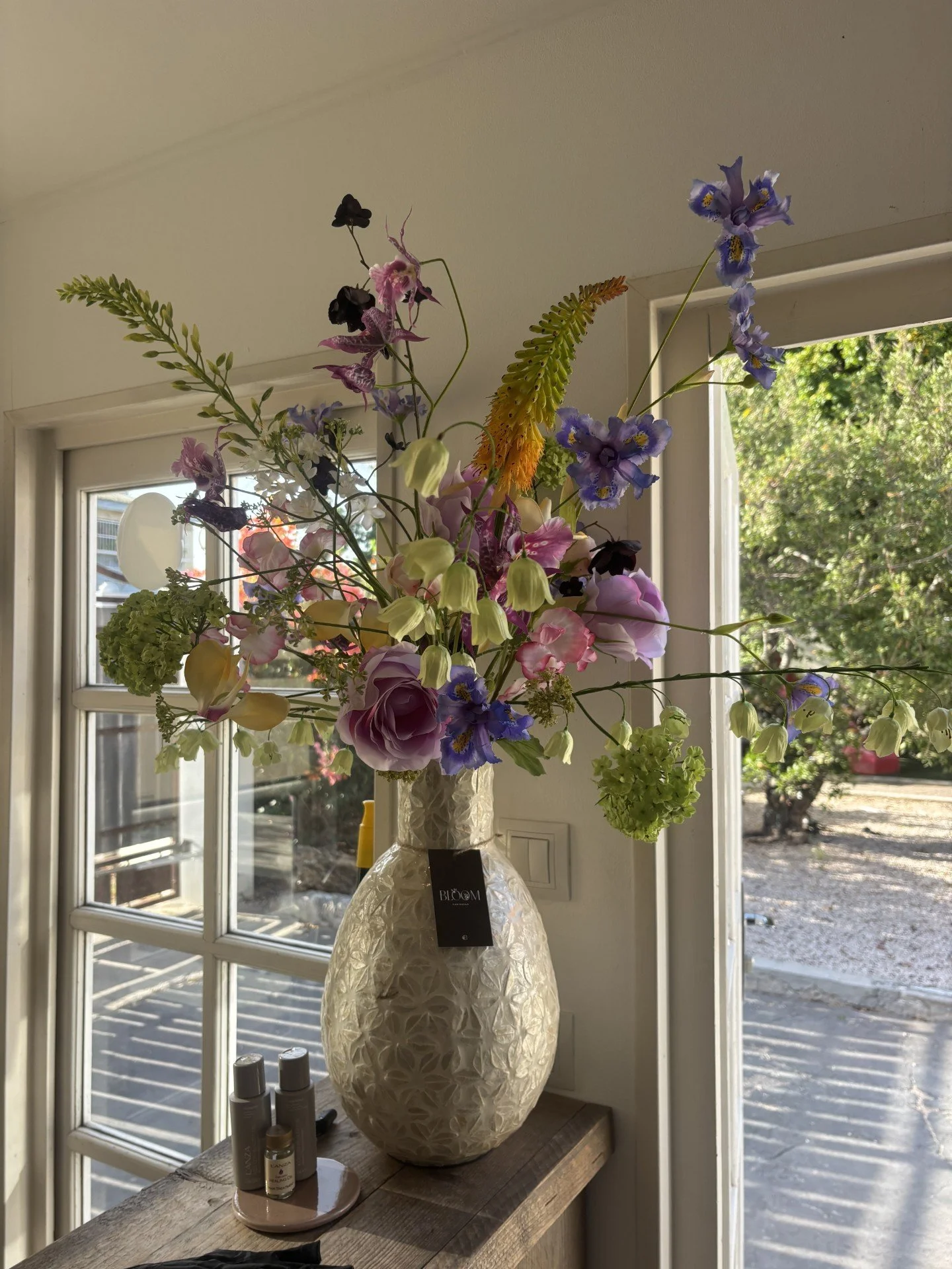 A floral arrangement in a textured white vase on a wooden table inside near a window.