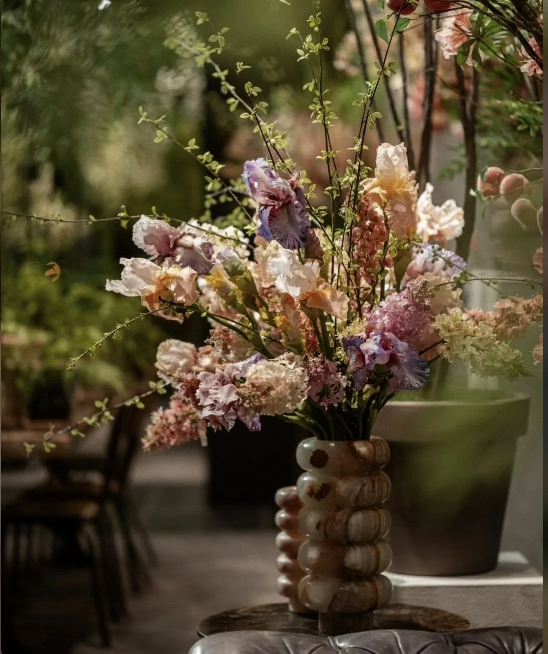 A tall ceramic vase with a textured, rounded design holds a bouquet of pink, purple, and cream flowers, with some greenery. The flowers are soft and ruffled, and the arrangement is placed on a table in a cozy indoor or outdoor setting, with potted plants and blurred background visible.