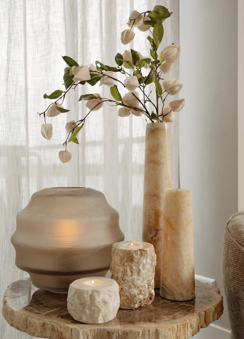 Decorative arrangement with a tall beige vase holding white flowers and green leaves, three candles of varying textures and sizes, and a large rounded beige vase on a wooden table in front of a sheer curtain.