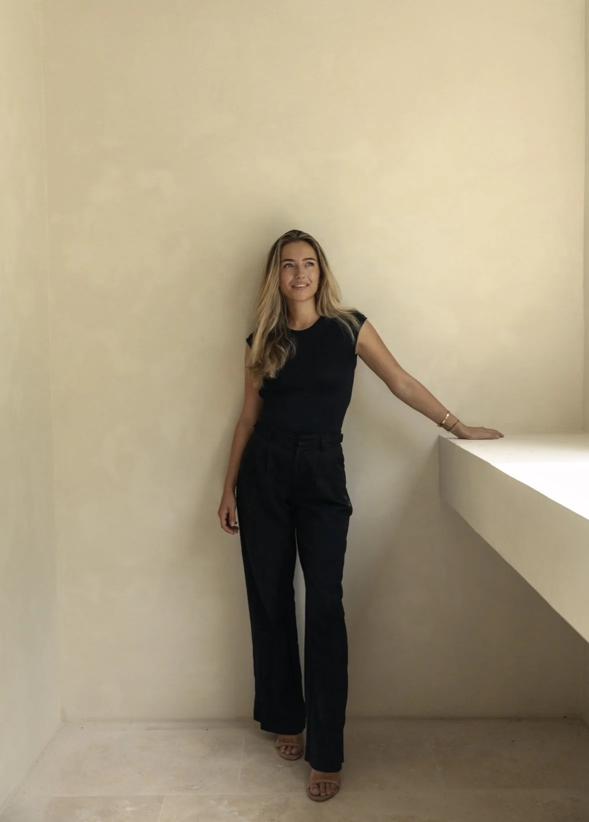 A woman with long blonde hair in a black top and black trousers standing with her left hand resting on a white ledge in front of a plain light-colored wall.