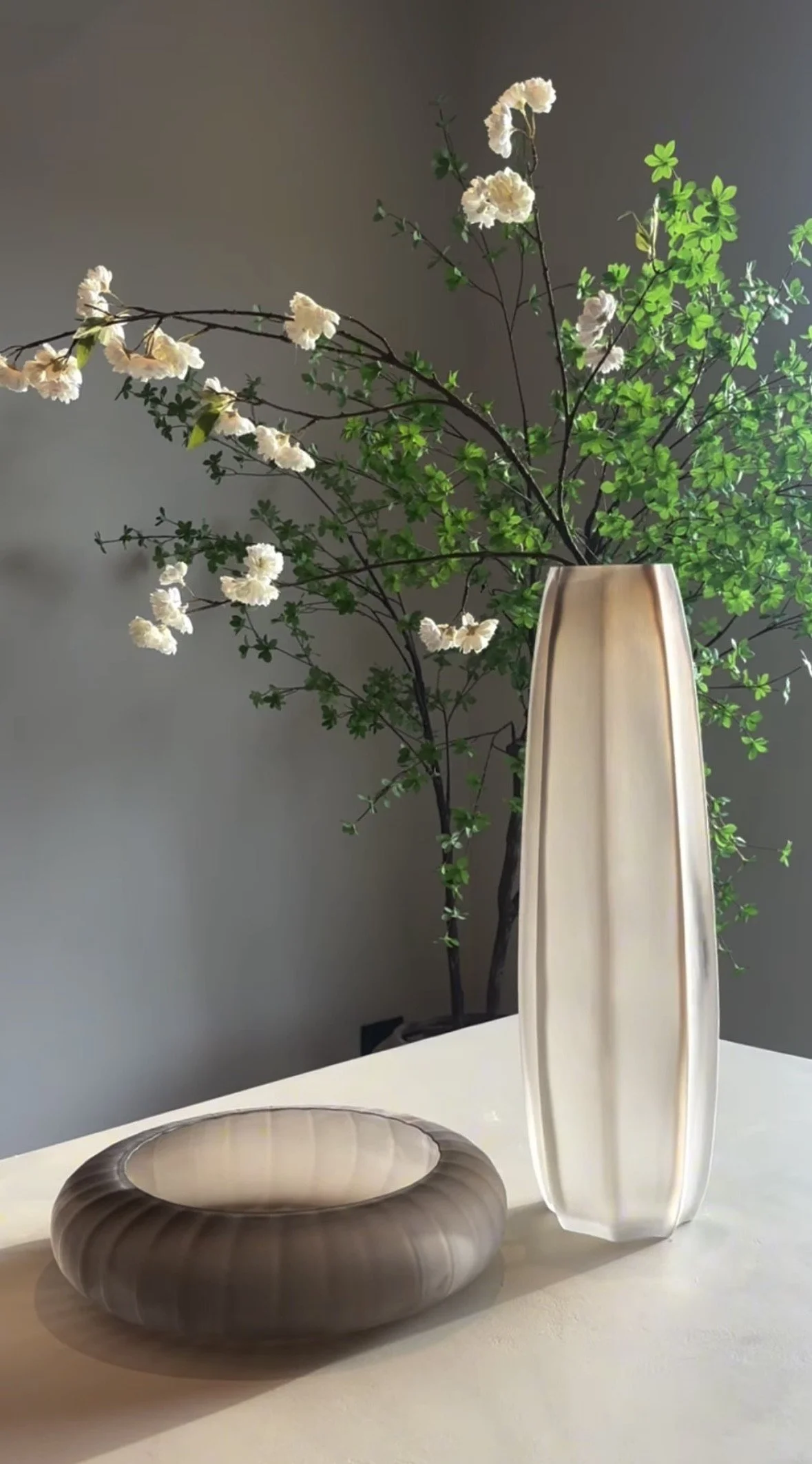 A tall, narrow beige and white vase with branches of white flowers and green leaves, placed on a table. A circular brown decorative object is on the table in the foreground.