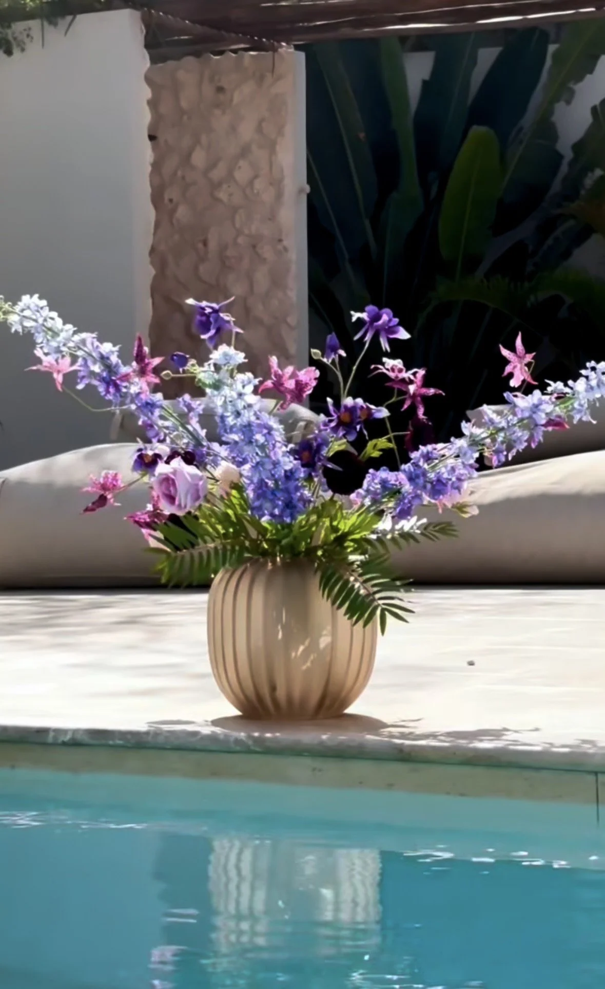 A beige ceramic vase with vertical ridges holds a bouquet of purple, pink, and white flowers, placed on a white surface by a pool.