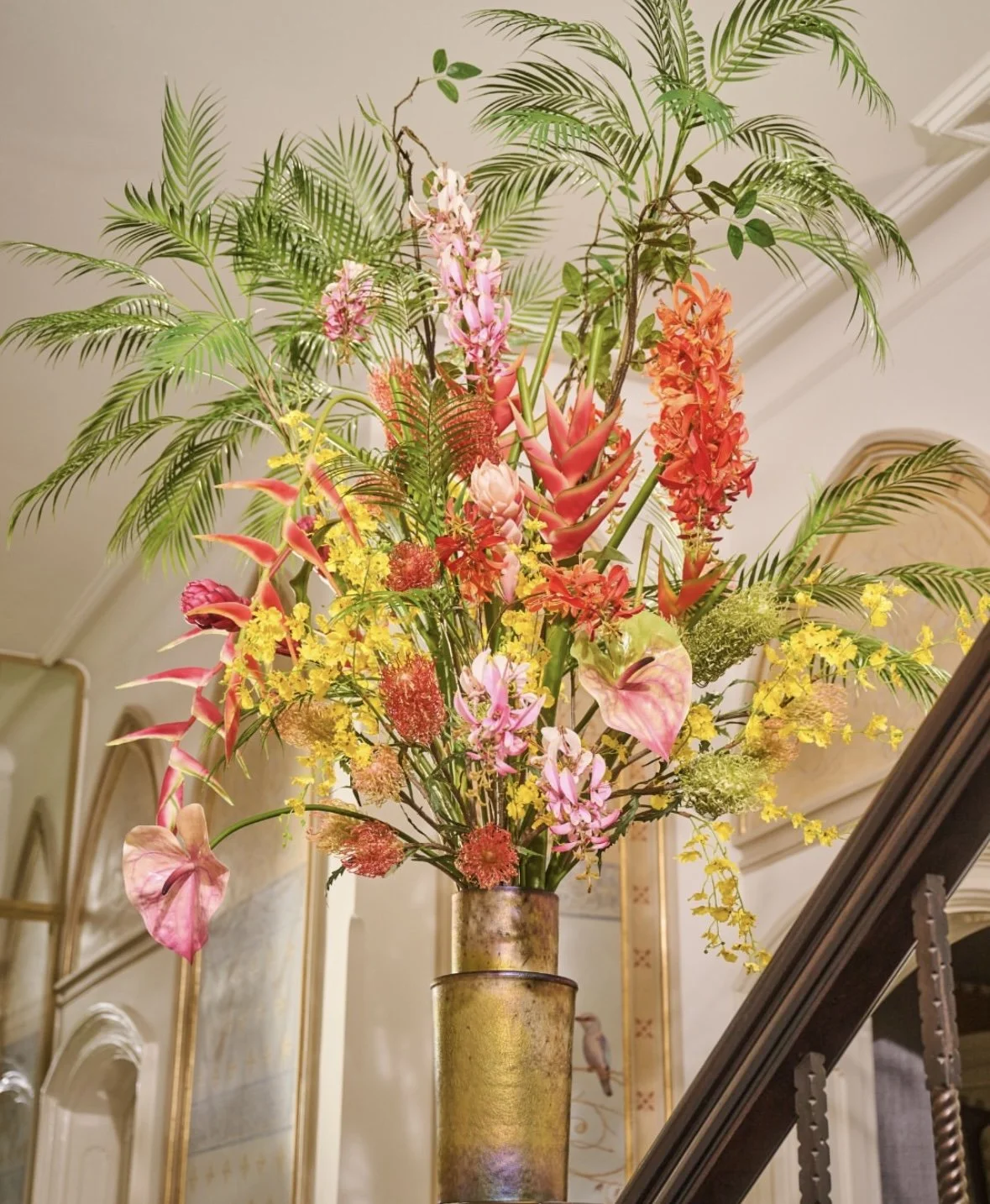 A large, colorful flower arrangement in a tall vase with various tropical and floral plants, including pink lilies, red orchids, yellow daisies, and green palm leaves, set against an elegant indoor background.