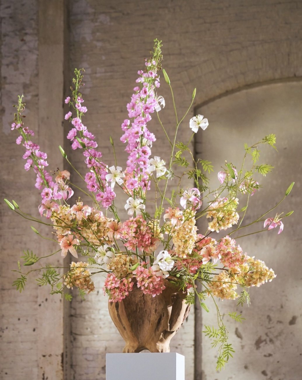 A bouquet of pink, white, and peach flowers arranged in a wooden vase, displayed on a pedestal against a brick wall.