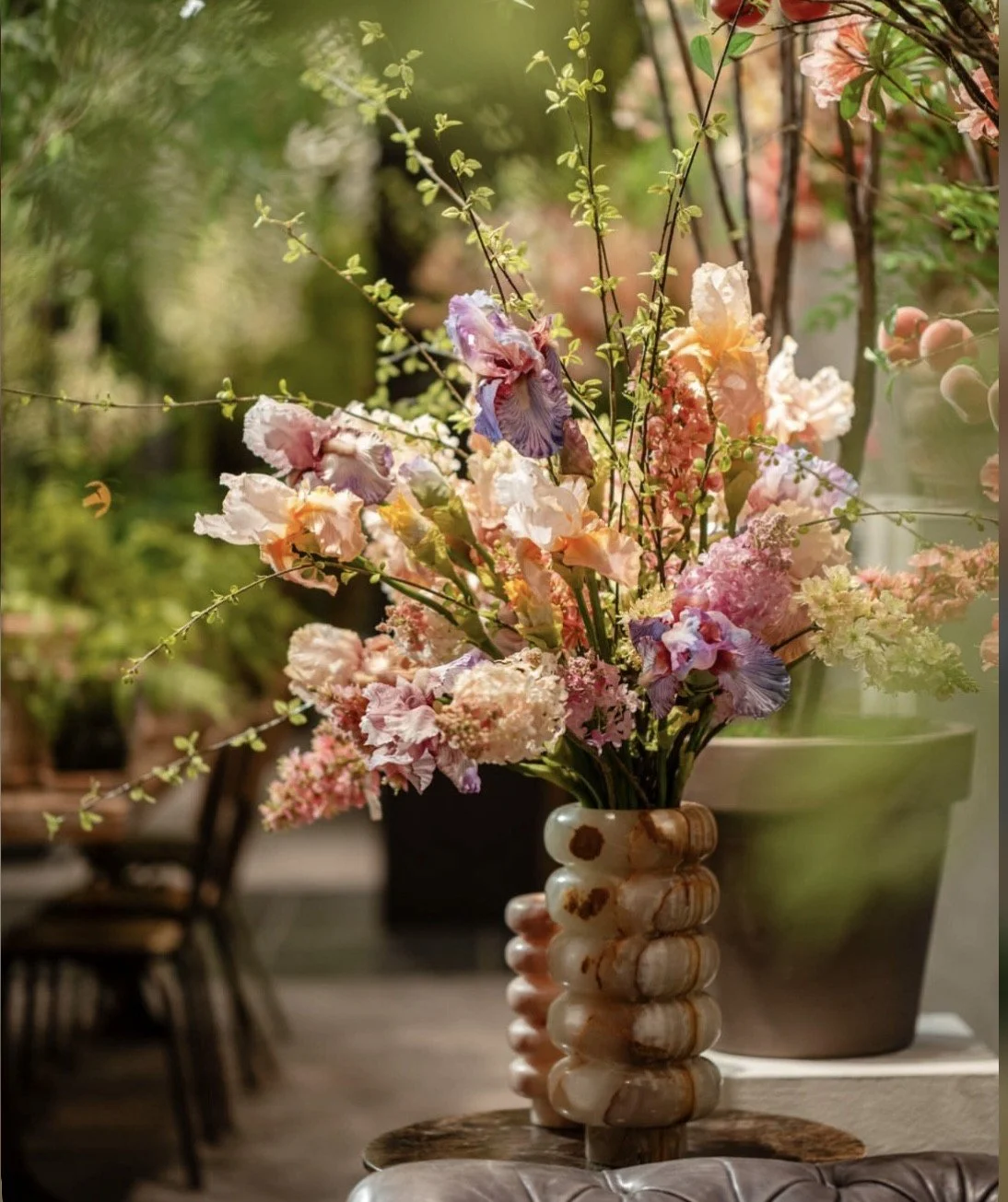 A bouquet of pink, white, and purple flowers arranged in a decorative beige and brown ceramic vase, placed on a wooden table with a blurred background of indoor and outdoor elements.