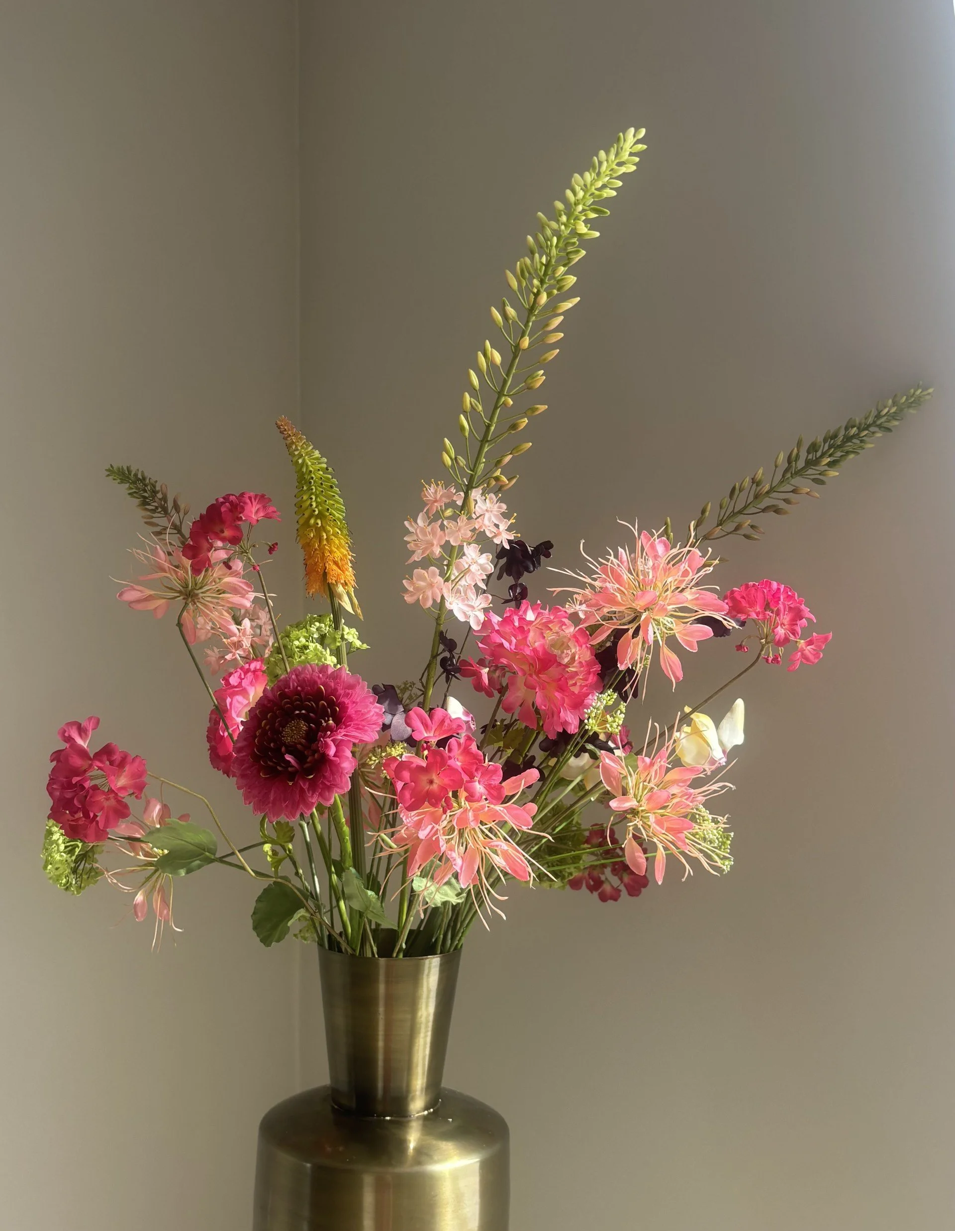 A tall bouquet of pink, green, yellow, and purple flowers in a shiny metallic vase on a dark round table against a gray wall.