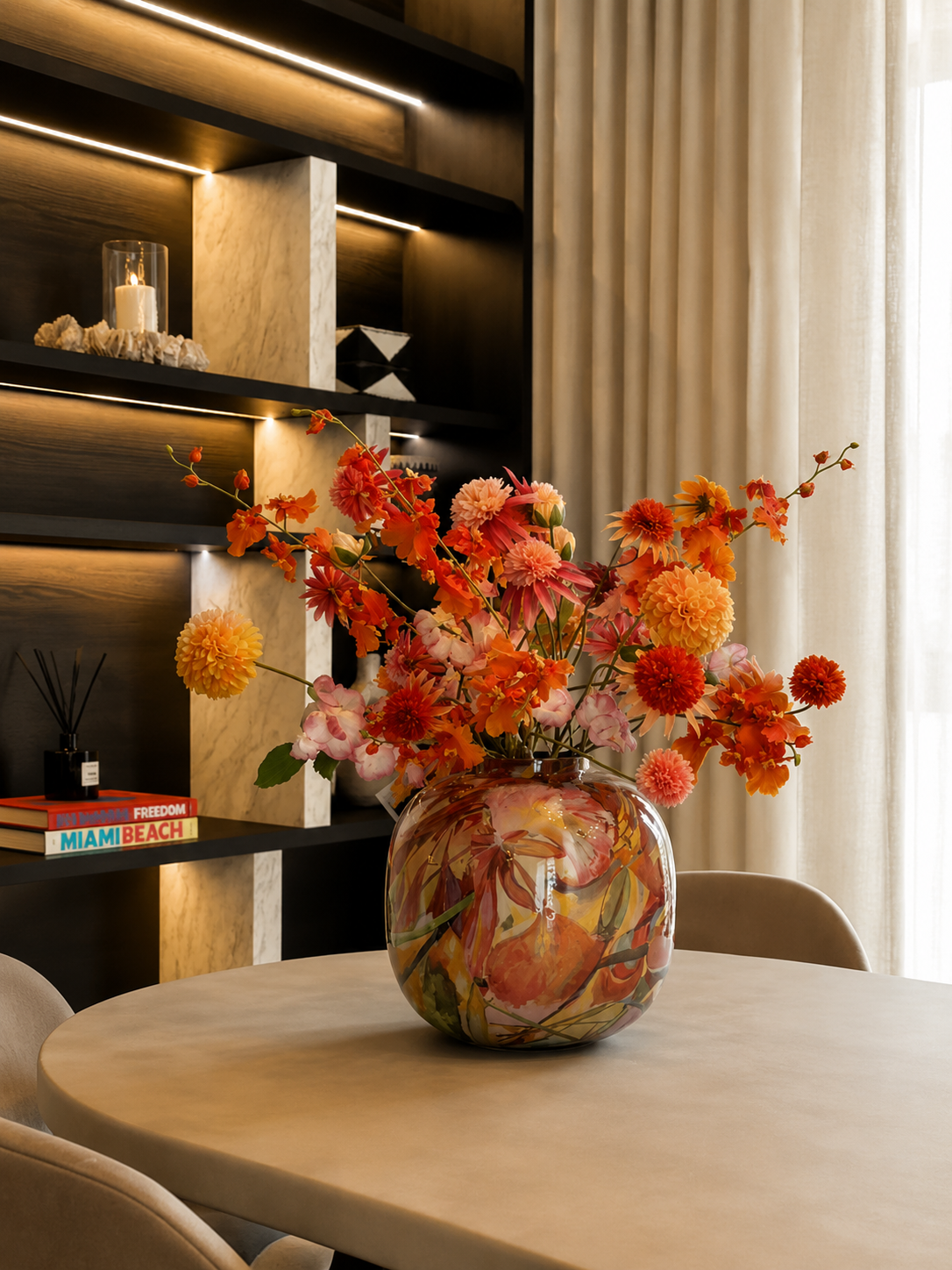 A round table with a colorful floral bouquet in a patterned glass vase, set in a modern living room with dark wood and marble shelving, books, and a candle, with light-colored curtains in the background.