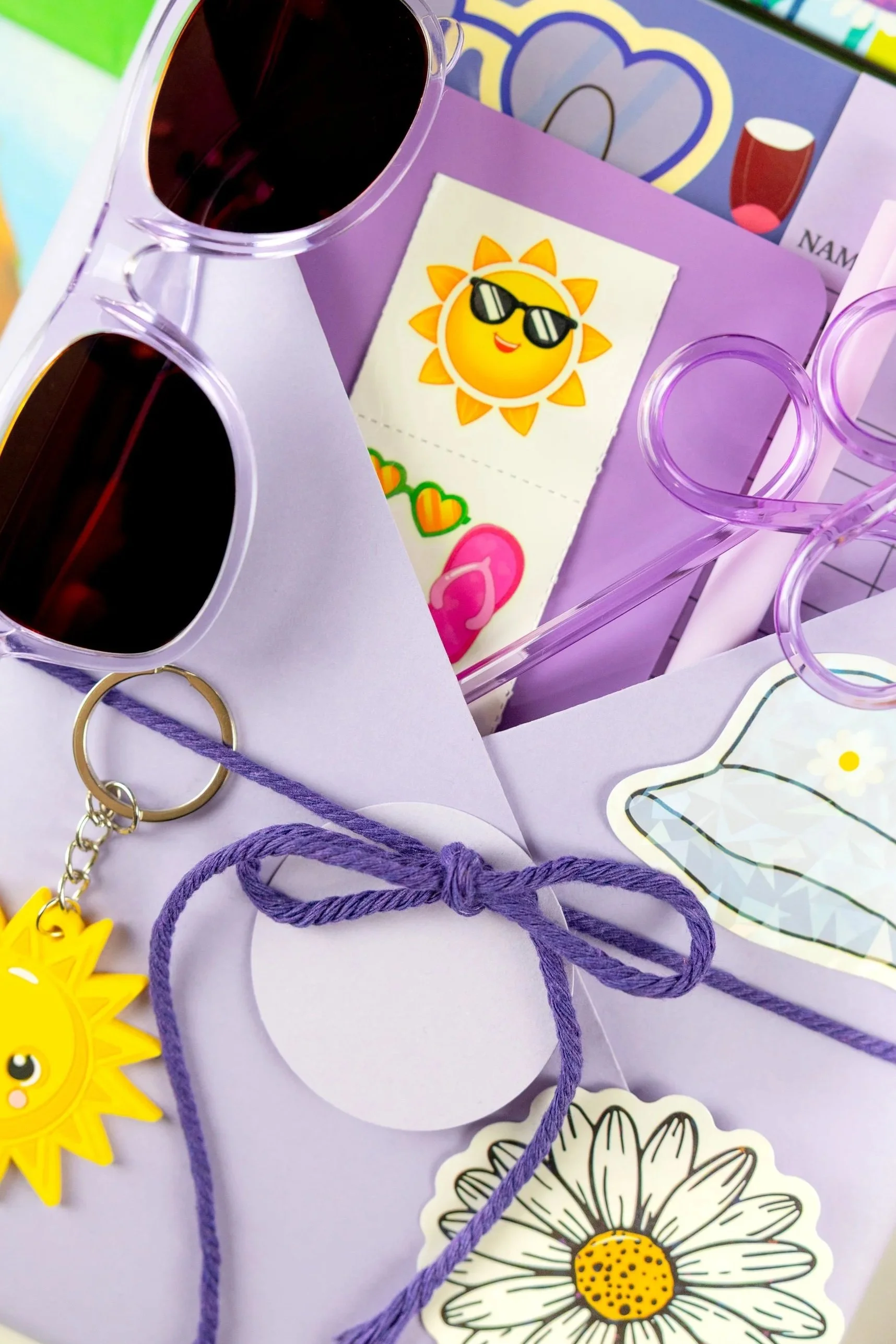 Close-up of colorful party-themed items, including sunglasses, cartoon sun with sunglasses, stickers of hearts and flowers, and purple string on pastel purple paper.