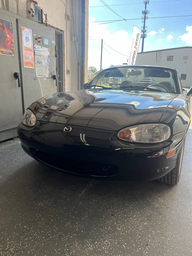 Black Mazda MX-5 Miata parked indoors with reflections on the hood, near a wall with posters and an outside view including sky and power lines.
