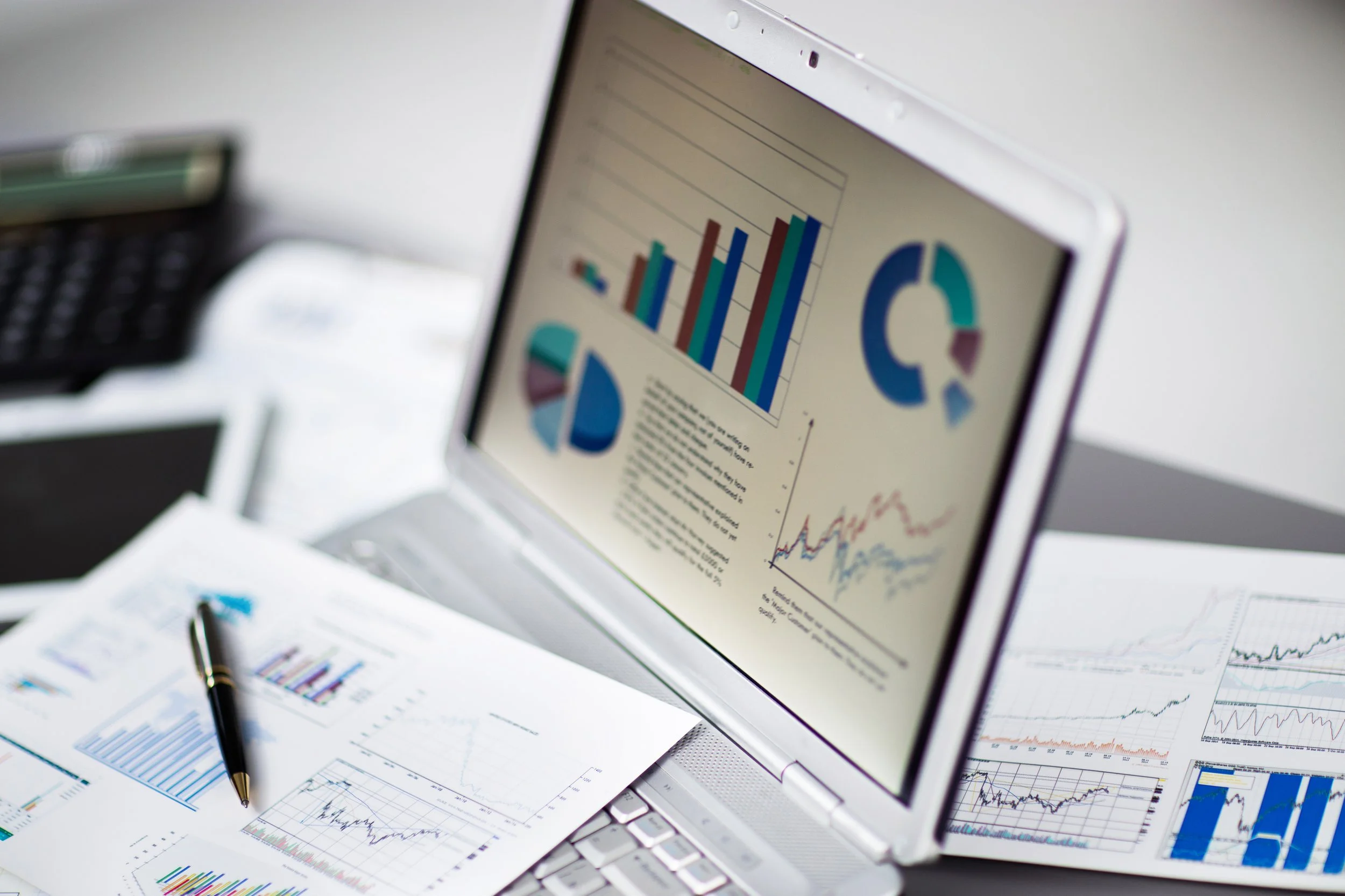 Laptop displaying various charts and graphs on the screen, with printed reports and a pen on the desk.