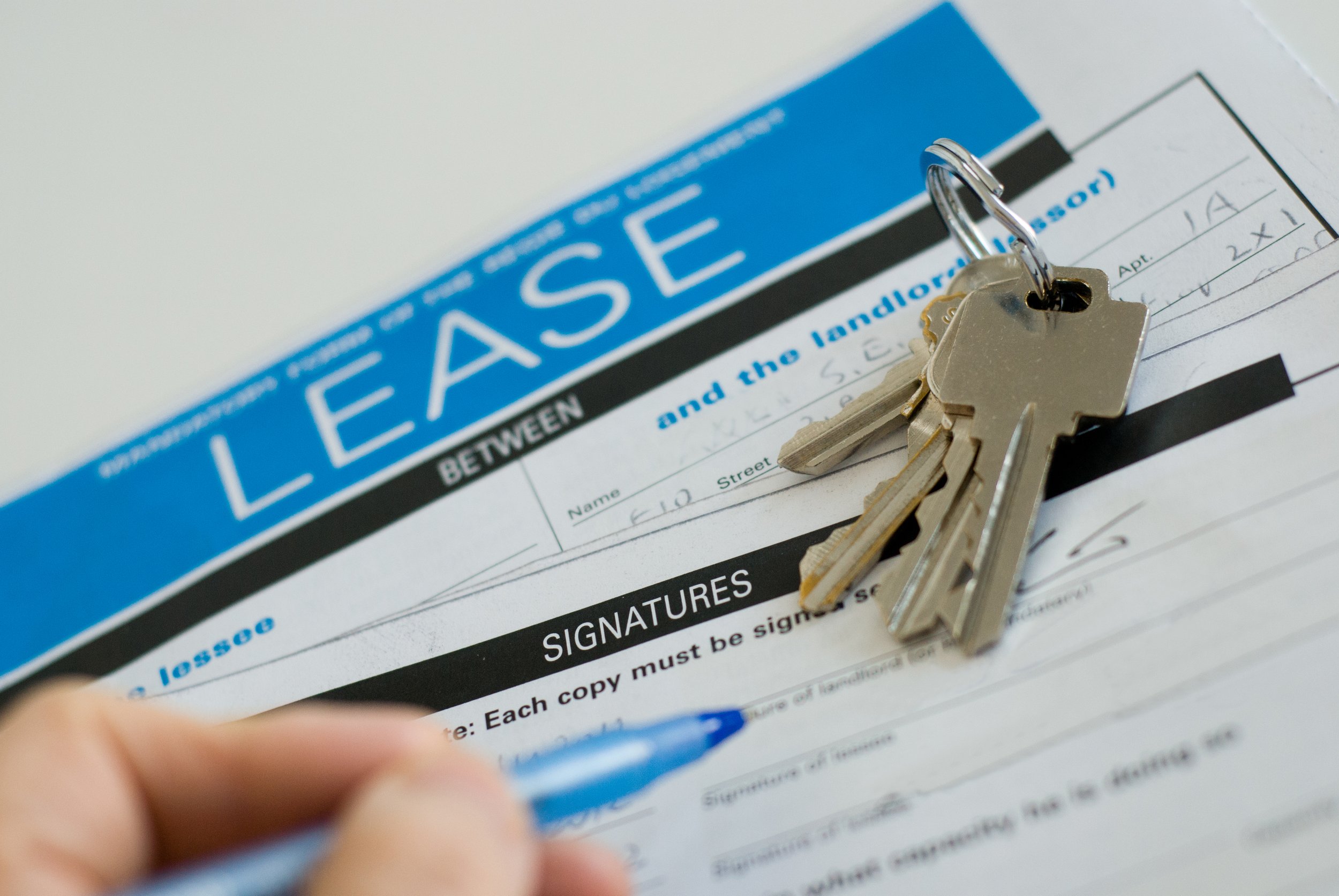 Close-up of a set of keys placed on a rental agreement form with a signature section, with a blue pen in a person's hand in the foreground.