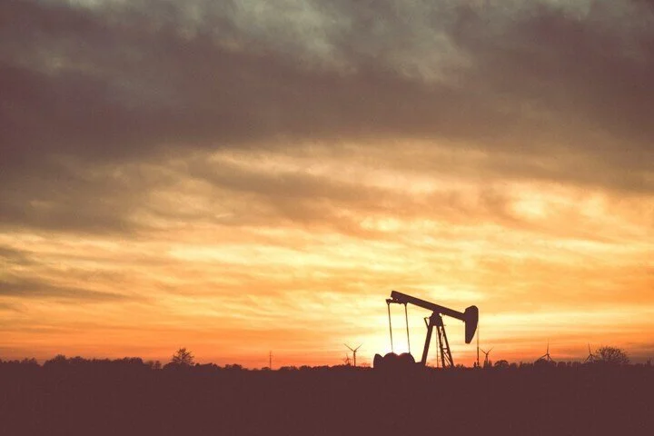 Oil pumpjack silhouetted against a colorful sunset sky with wind turbines in the distance.