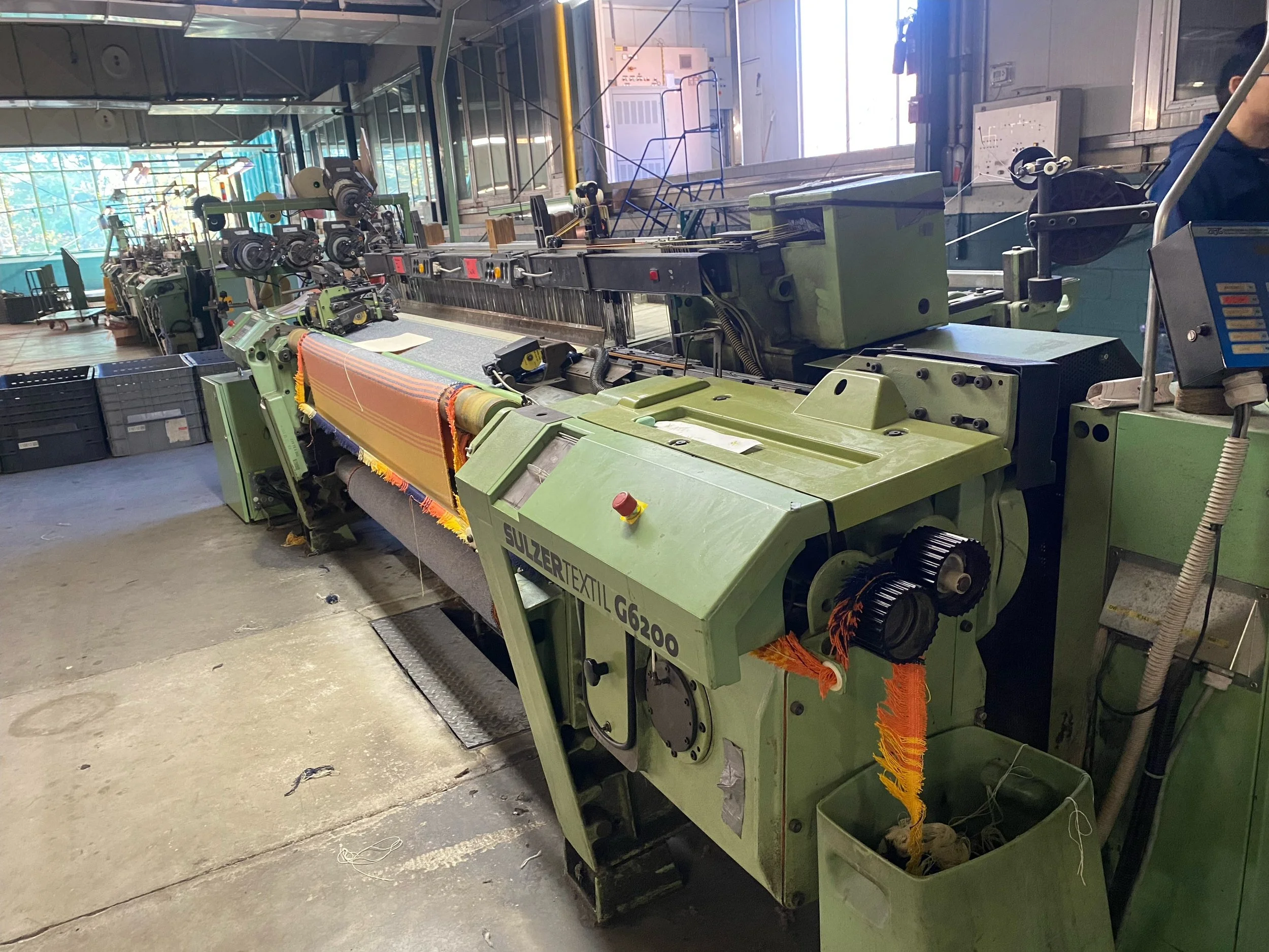Inside an industrial textile manufacturing facility, a large green textile machine is in operation, with fabric being woven or dyed. The machine has various controls and rollers, and there are some open containers and tools nearby, with sunlight coming through large windows in the background.