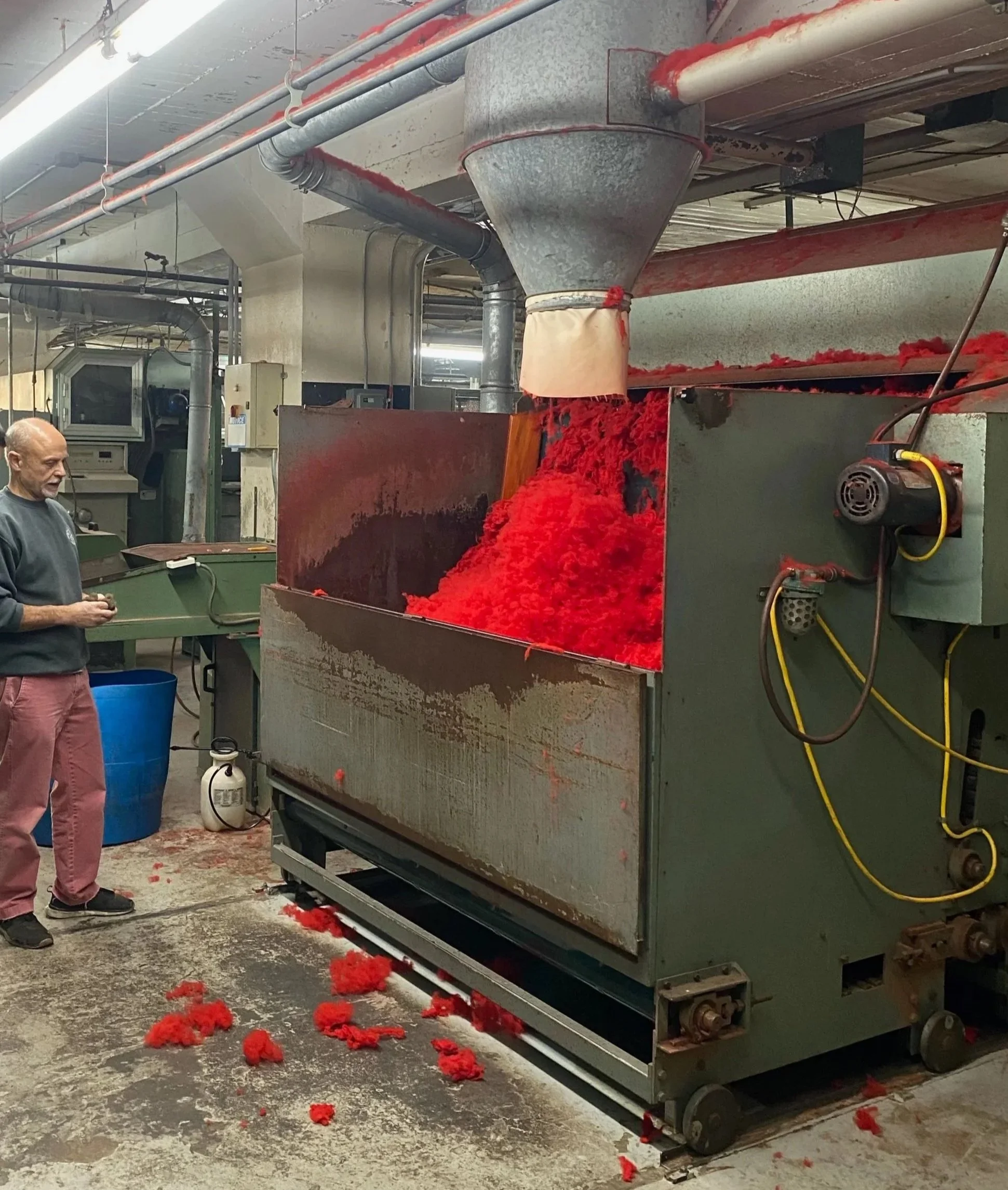 An industrial machine is processing red fiber material, with scattered bits on the floor around it. A person is standing nearby, observing or operating the equipment in a workshop or factory setting.