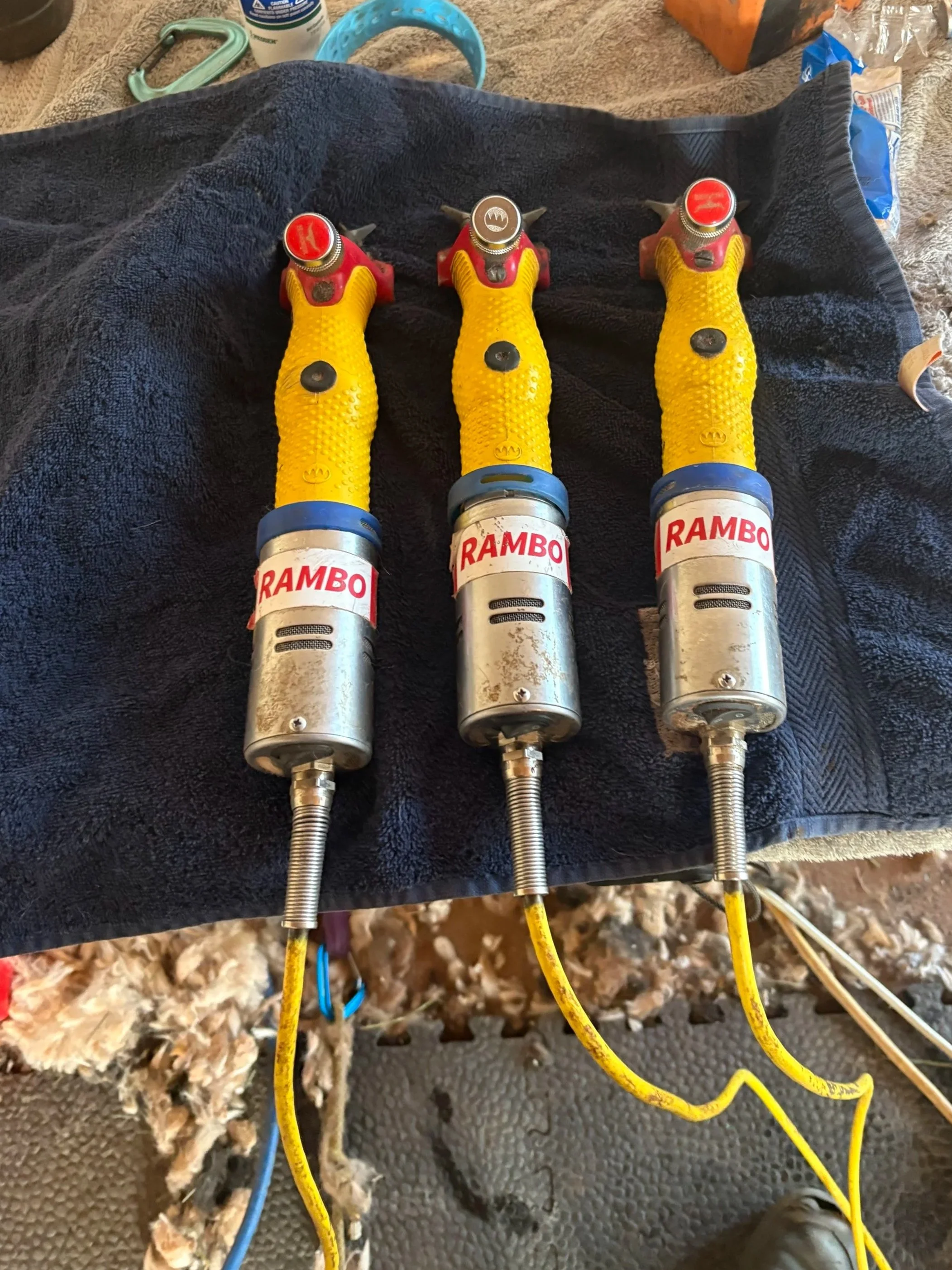 Close-up of three professional shearing machines with yellow handles, labeled 'RAMBO', connected by yellow and blue cords on a textured black surface. Surrounding debris, alpaca fiber, and a work environment background.