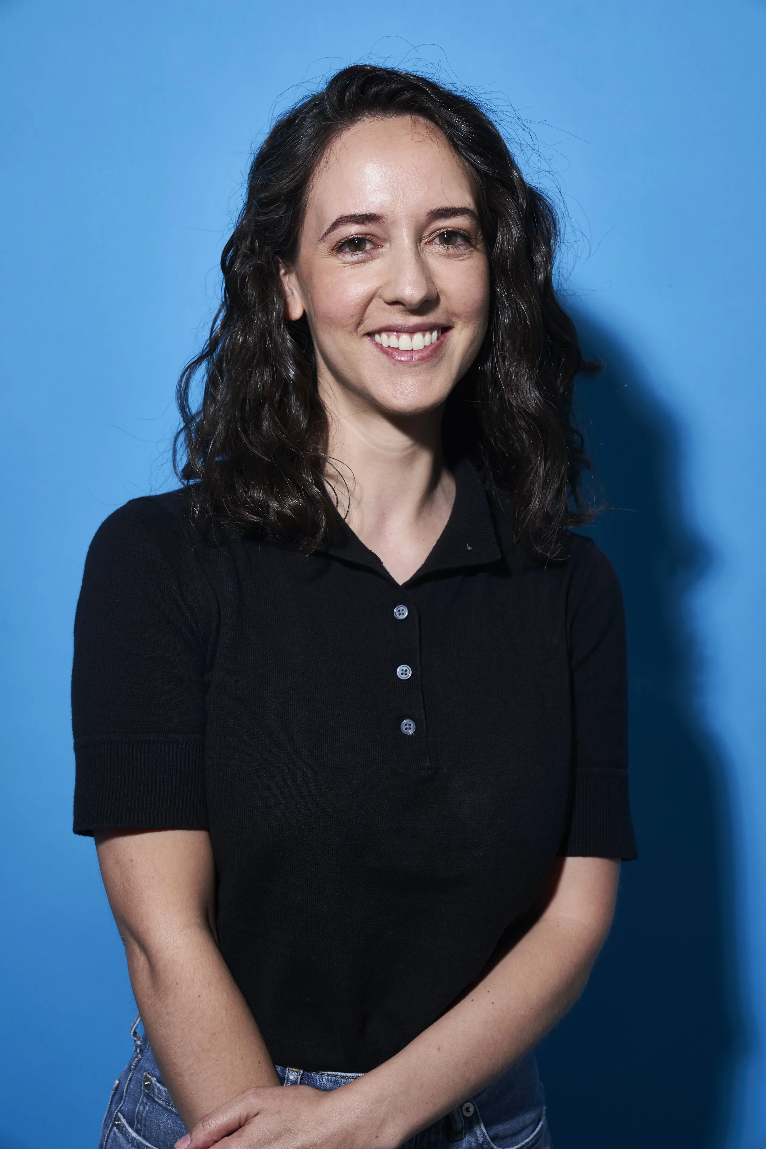 A smiling woman with dark hair wearing a white button-up shirt and light gray pants, sitting with a closed MacBook laptop on her lap against a black background.