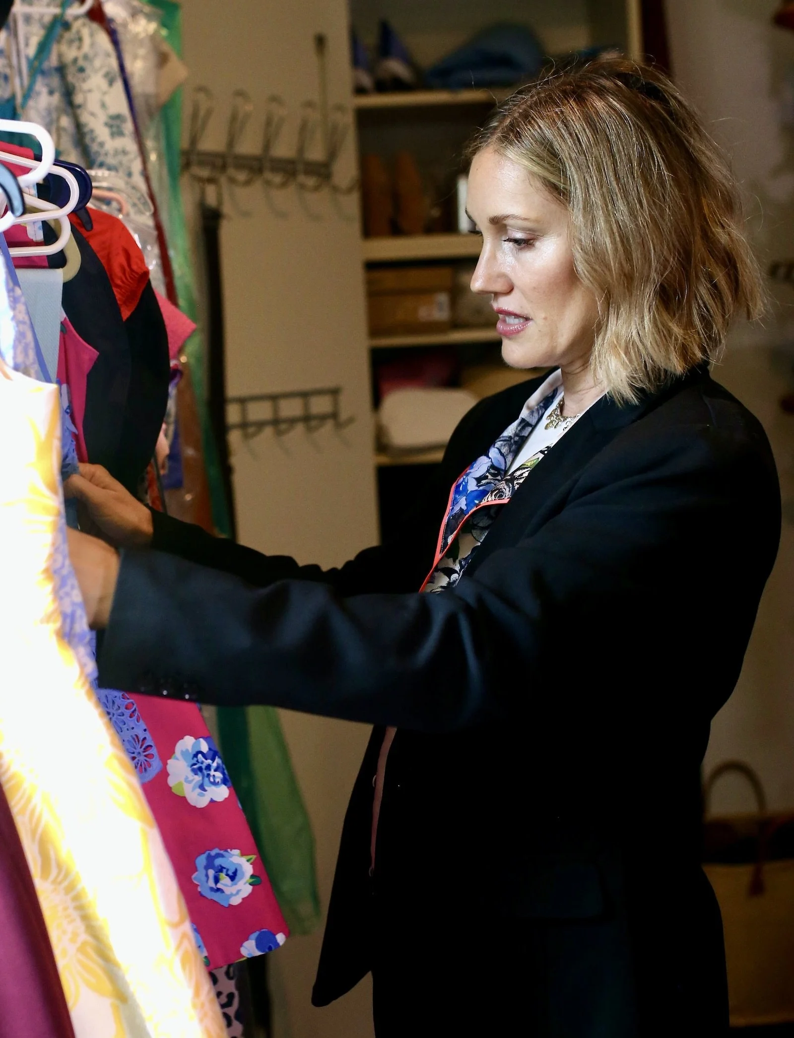 Fashion stylist and designer Carissa Macedo looks through a rack of clothes during a closet styling and organization session. She's wearing a black blazer and has short blonde hair.