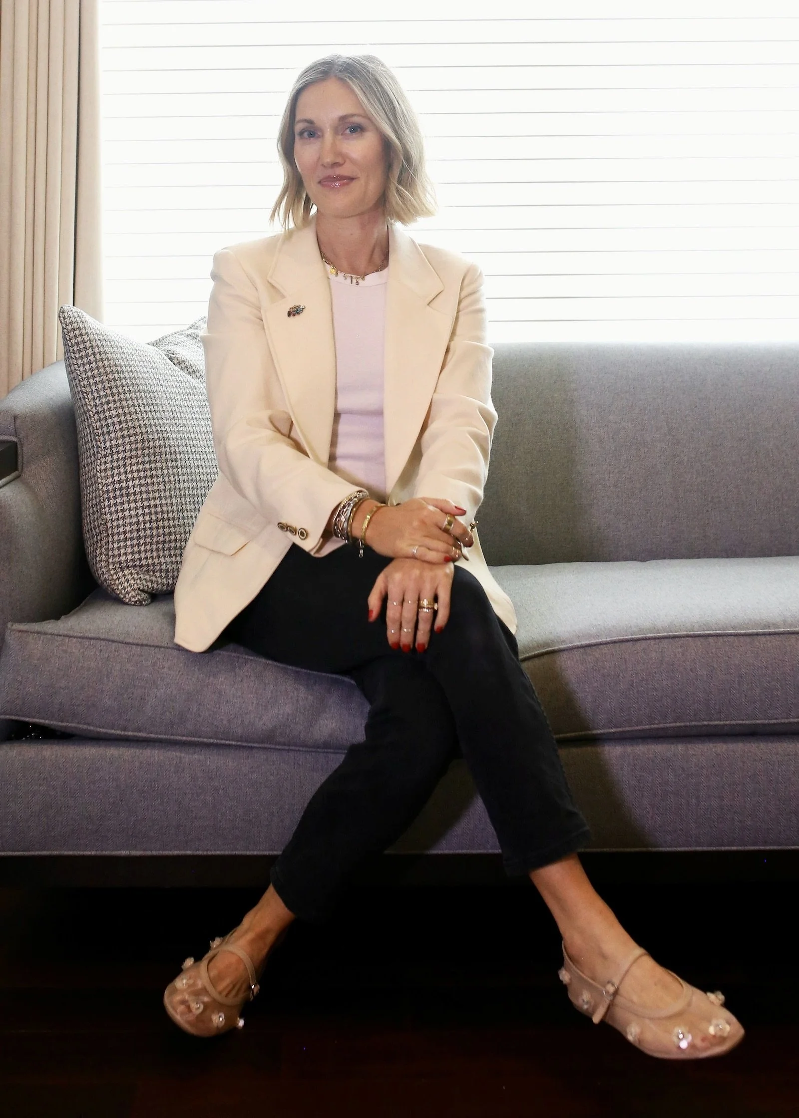 Fashion stylist and designer Carissa Macedo sits on a gray couch in front of a window. She's wearing an off-white blazer and black pants with beige see-through flats. She has short blonde hair and is smiling.