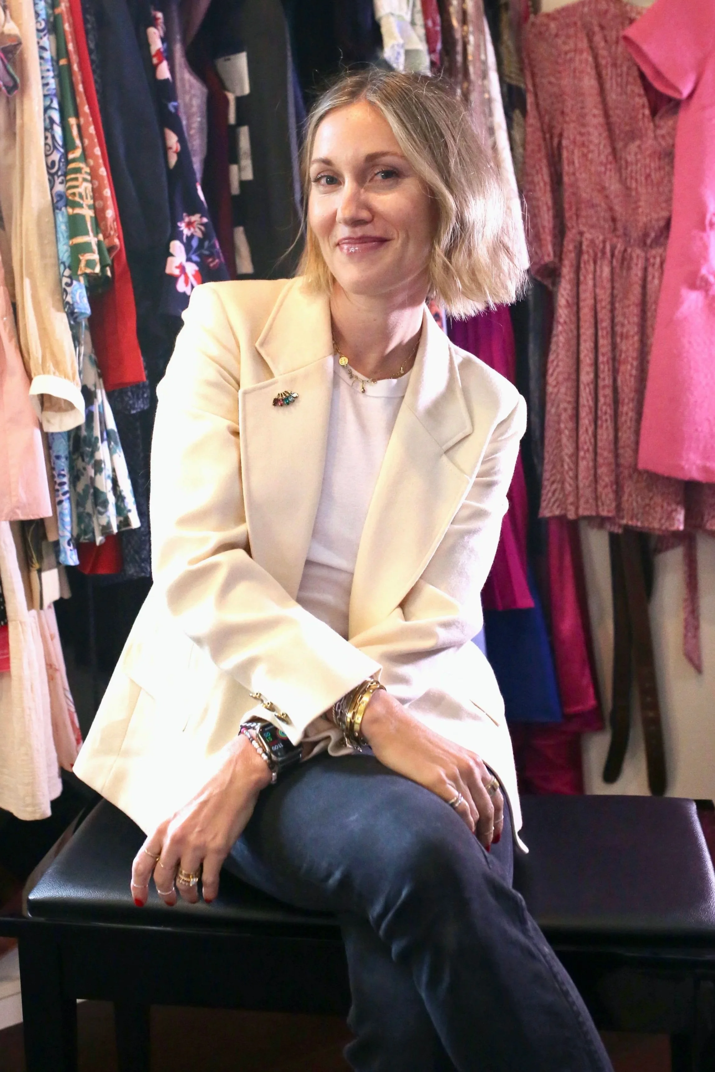 Fashion stylist and designer Carissa Macedo sits in a closet with colorful clothes in the background. She's wearing a white blazer with a jeweled broach and jeans. She has short blonde hair and is smiling.