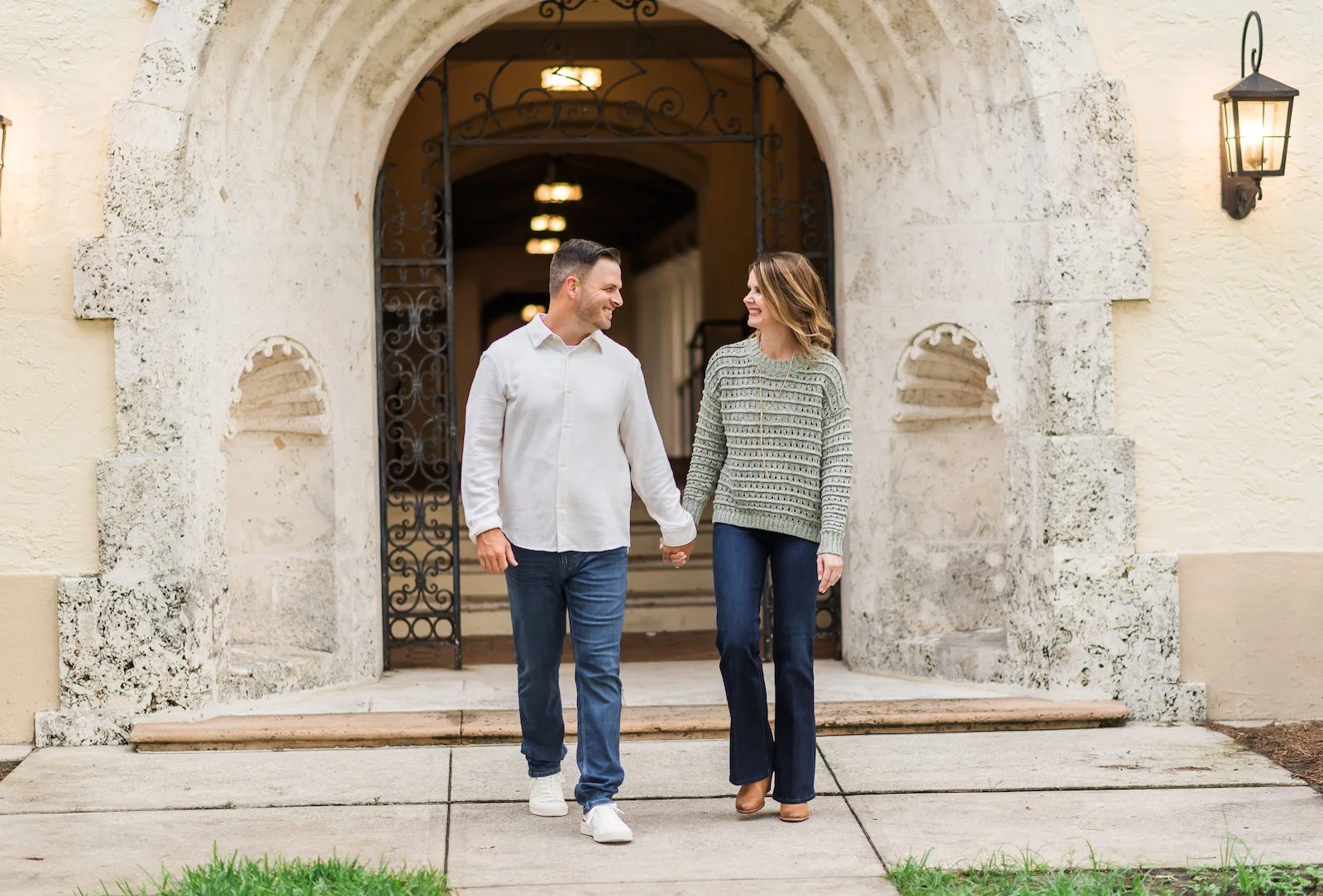 A person wearing a white collared shirt and jeans holds hands with a person wearing a green sweater and jeans while walking in front of an archway during a photo shoot.