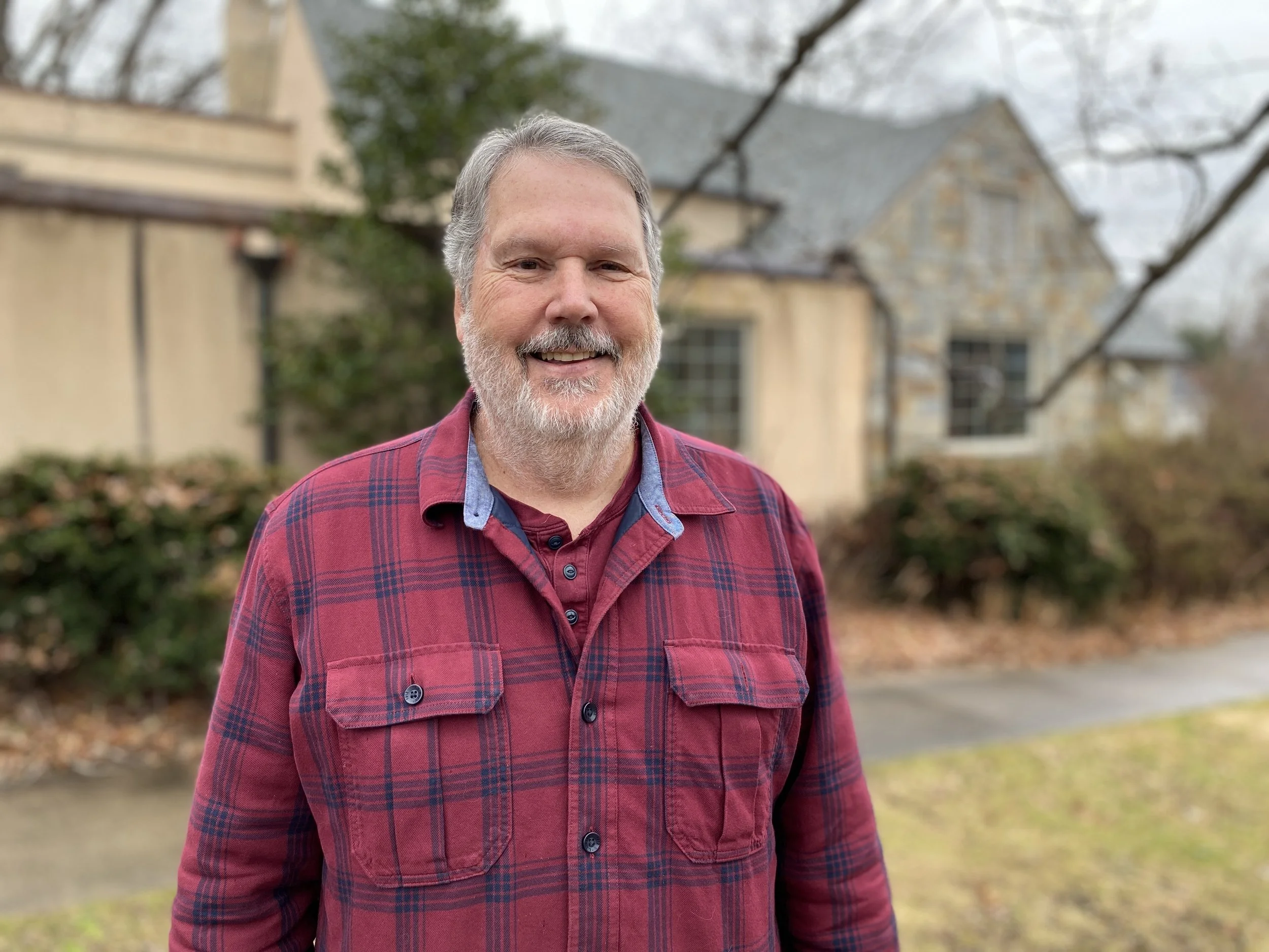 A smiling middle-aged man with gray hair and a beard standing outdoors in front of houses and trees on a cloudy day.