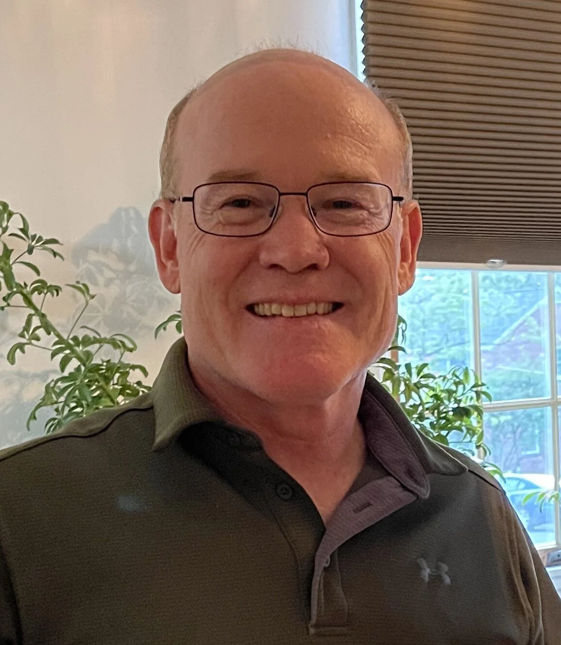 A middle-aged man smiling, wearing glasses and a dark collared shirt, indoors with a window and plants in the background.