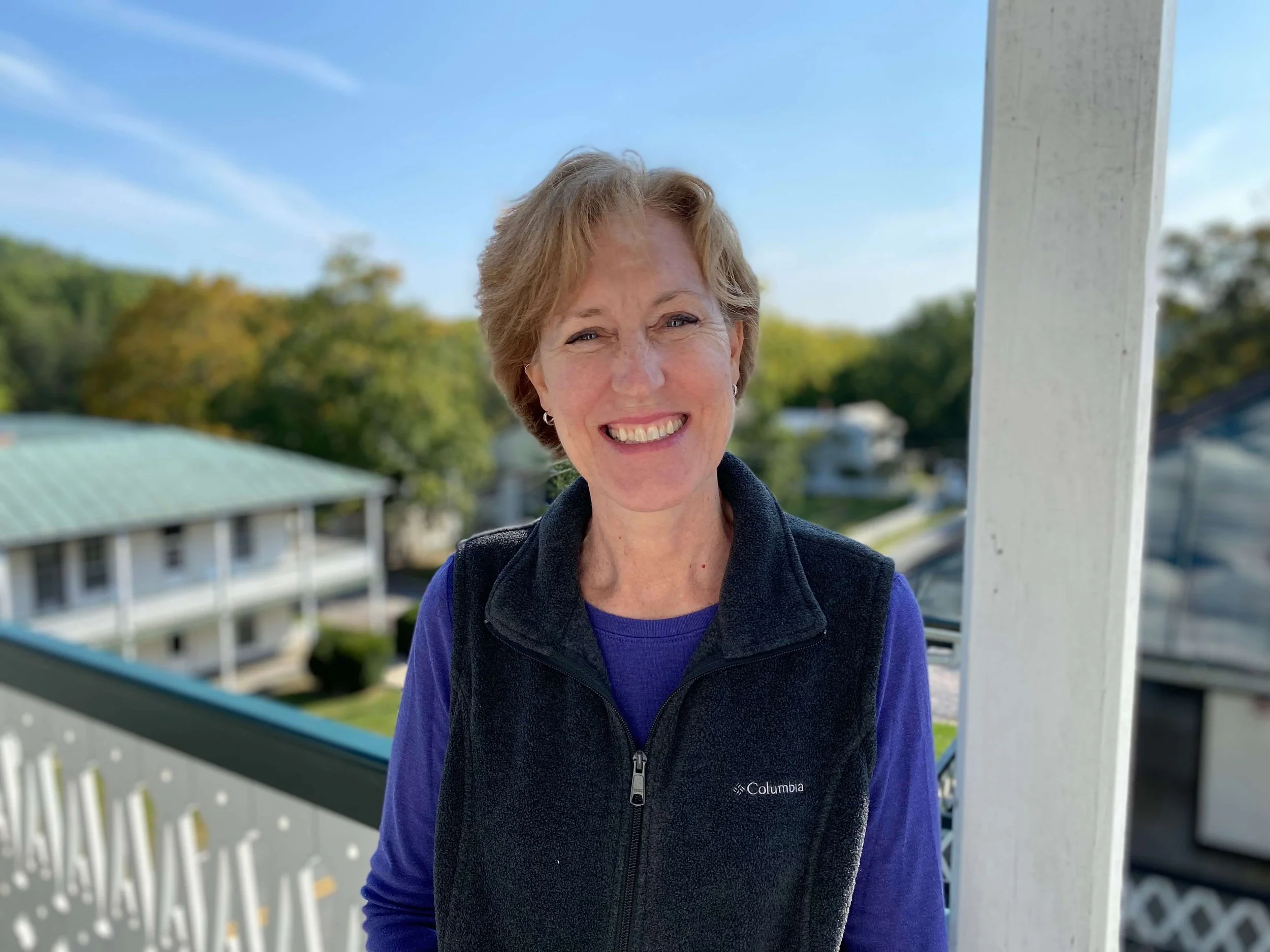 Smiling woman with short hair standing outdoors on a balcony with trees and houses in the background, wearing a black Columbia vest over a purple shirt.