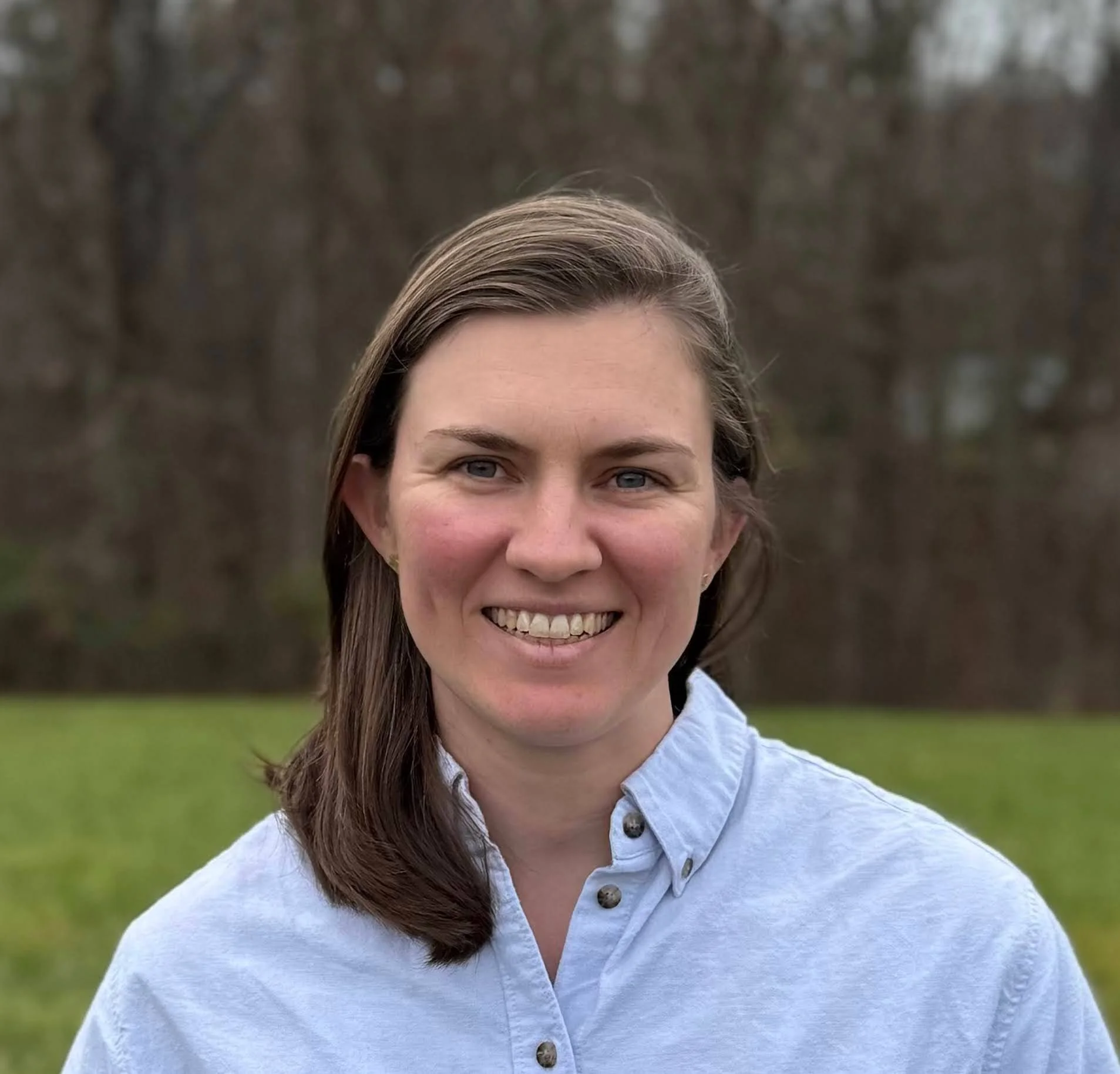 Portrait of a woman with brown hair, smiling, wearing a light blue shirt, standing outdoors with a blurred background of trees and grass.
