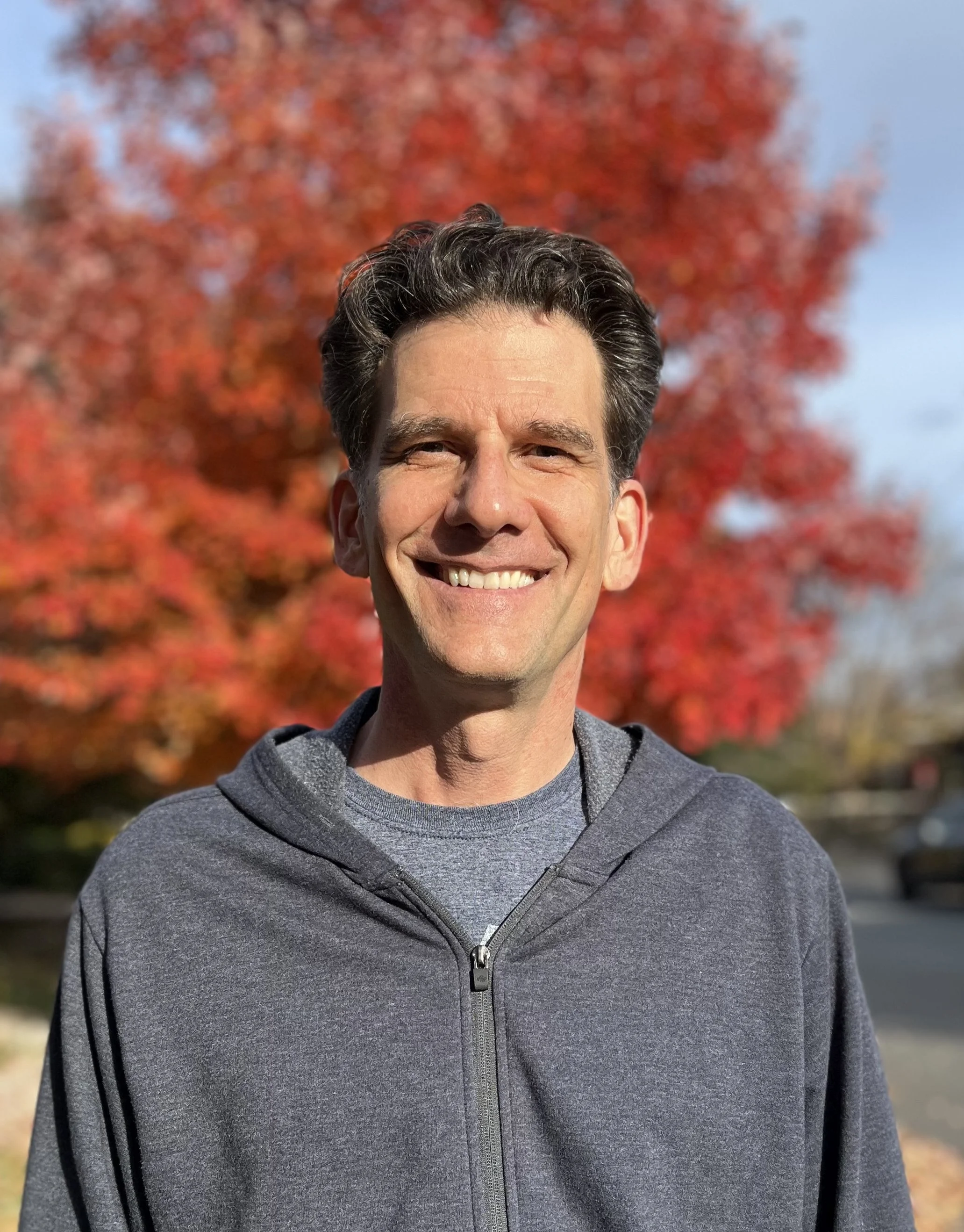 A smiling man with dark, wavy hair in a gray zip-up hoodie, standing outdoors with a vibrant red autumn tree in the background on a sunny day.